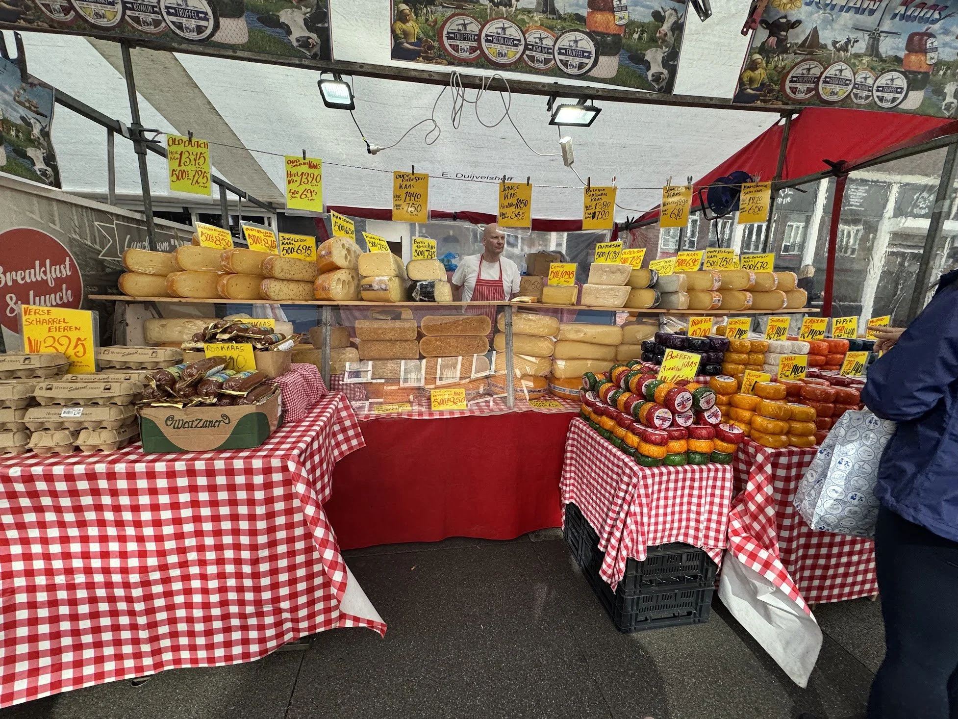 A market stall with various types of cheese on display, all labeled with yellow and orange signs indicating prices and names. The stand has a red and white checkered tablecloth, and a man wearing a white shirt and red apron is behind the counter. The