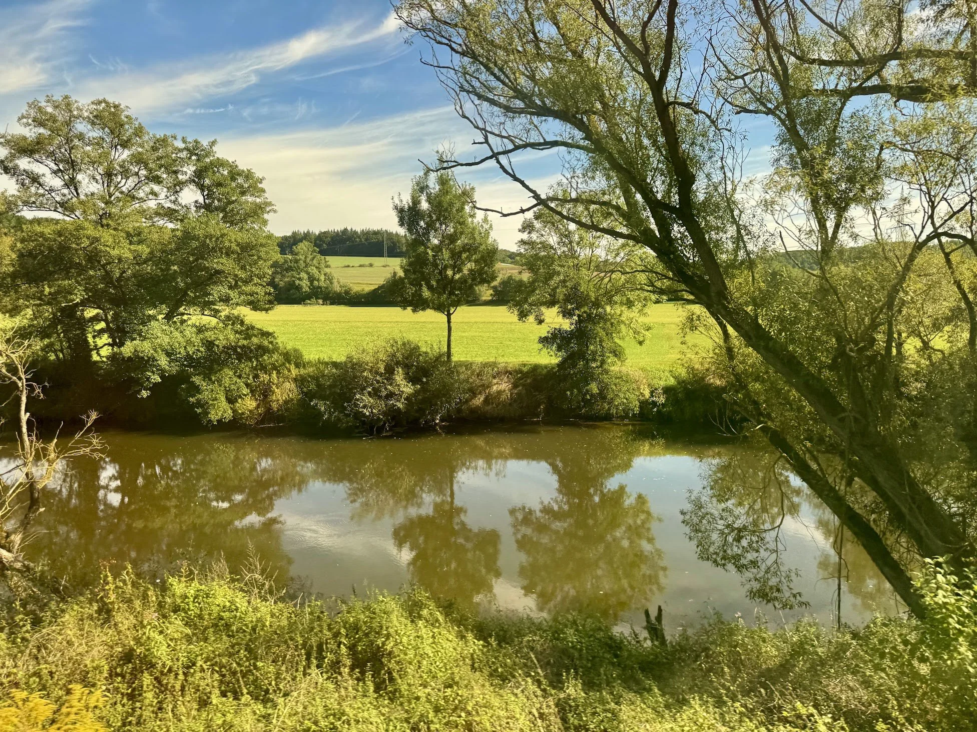 A peaceful landscape featuring a calm river with reflections of trees, a grassy field, and a blue sky with wispy clouds.