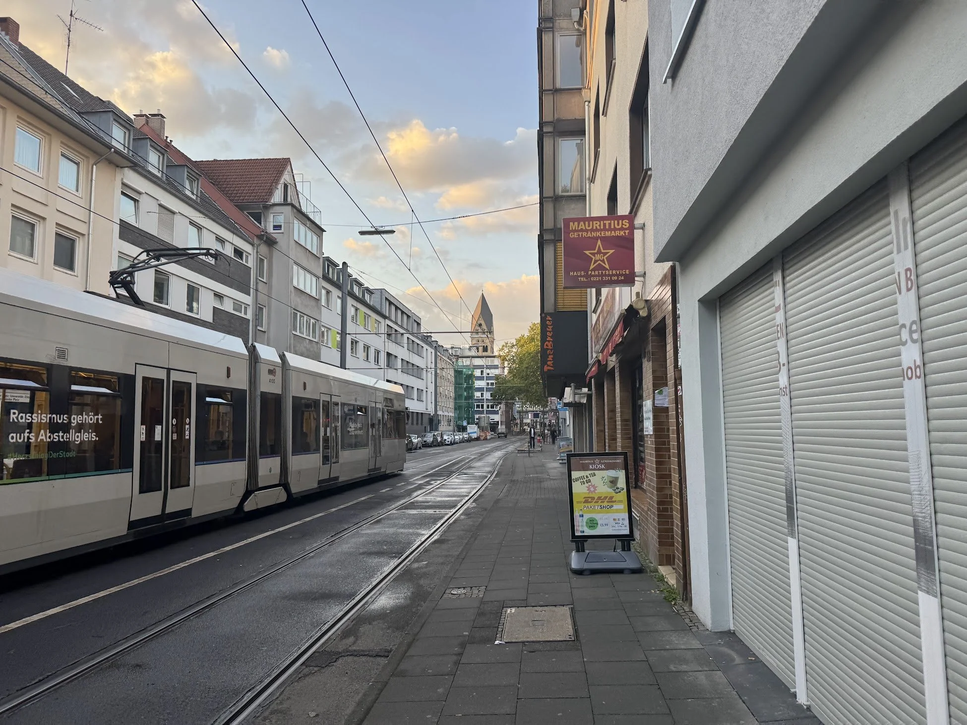 City street scene with tram on tracks, overhead wires, multi-story buildings, a church steeple in the distance, and a sidewalk with signs and storefronts.
