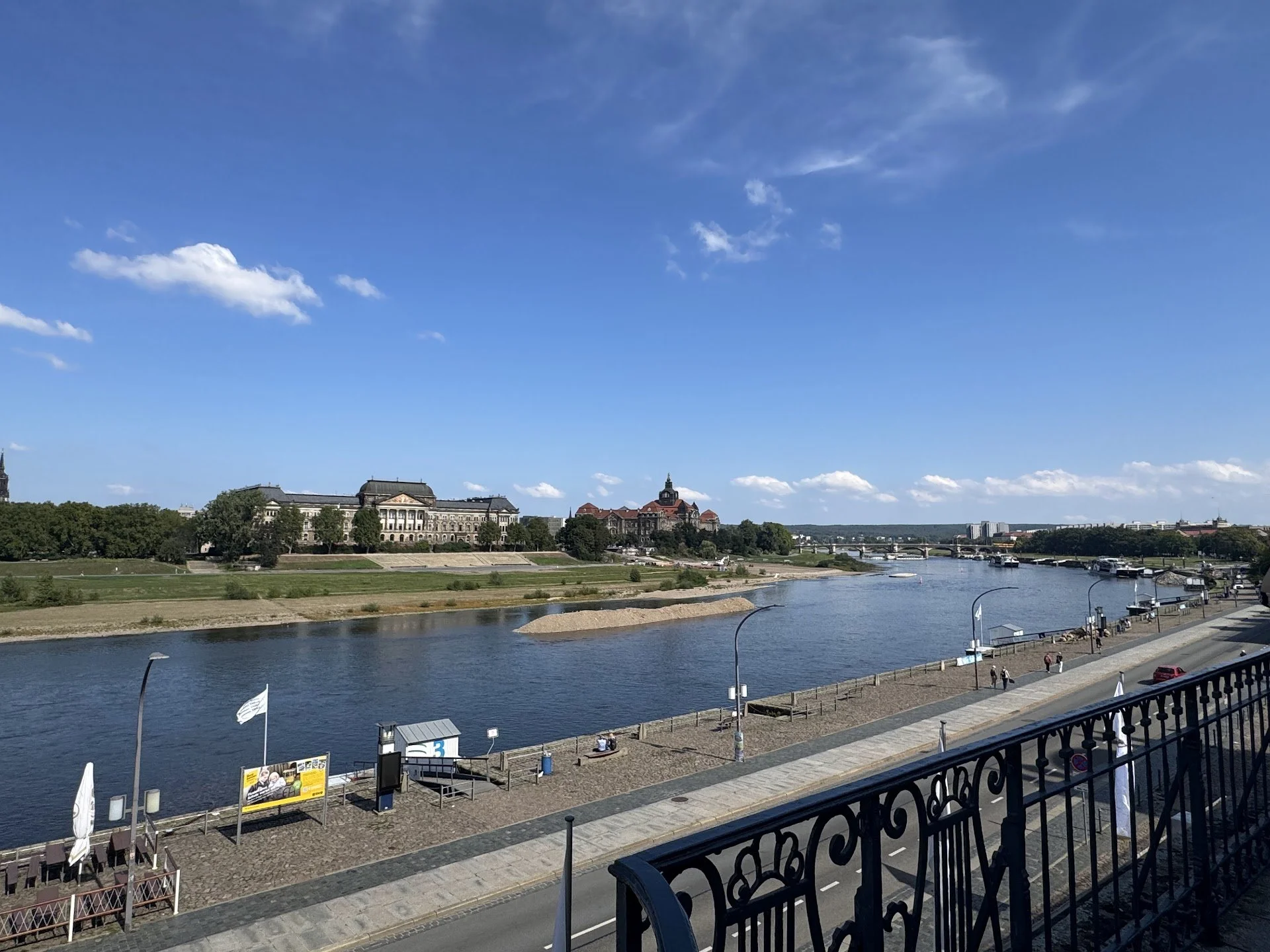 Cityscape view of a river with a historic building, bridge, and trees under a blue sky with some clouds.
