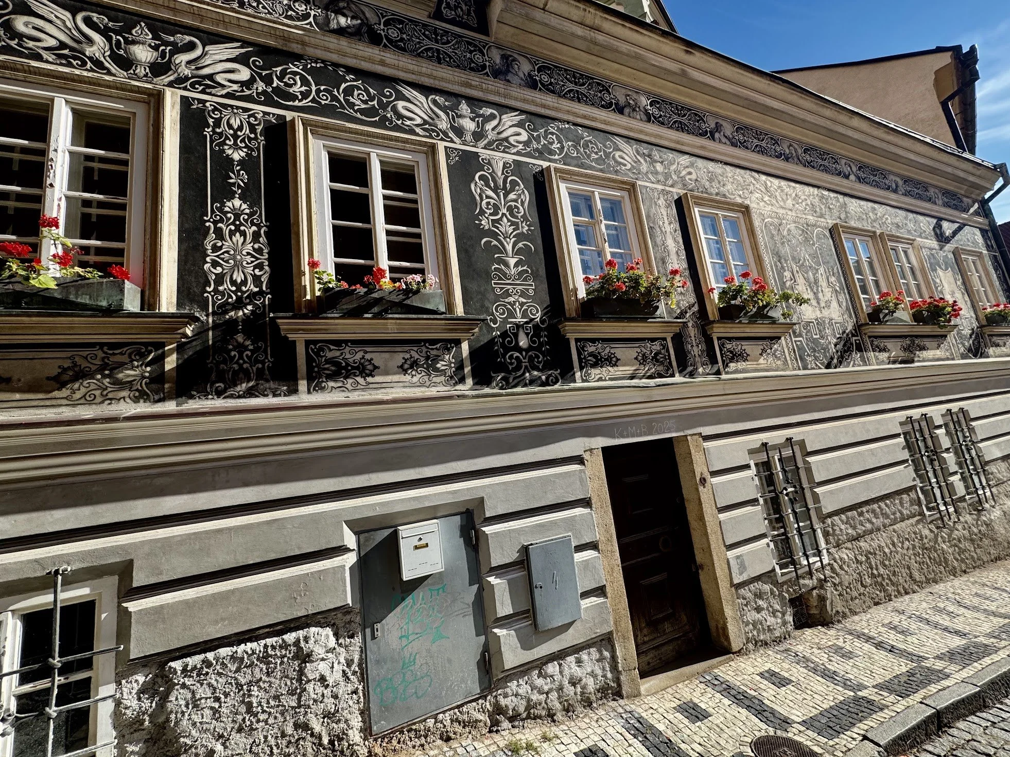 A historic building with decorative black and white painted designs on its facade, featuring multiple windows with flower boxes of red flowers, and a stone base with a sidewalk in front.