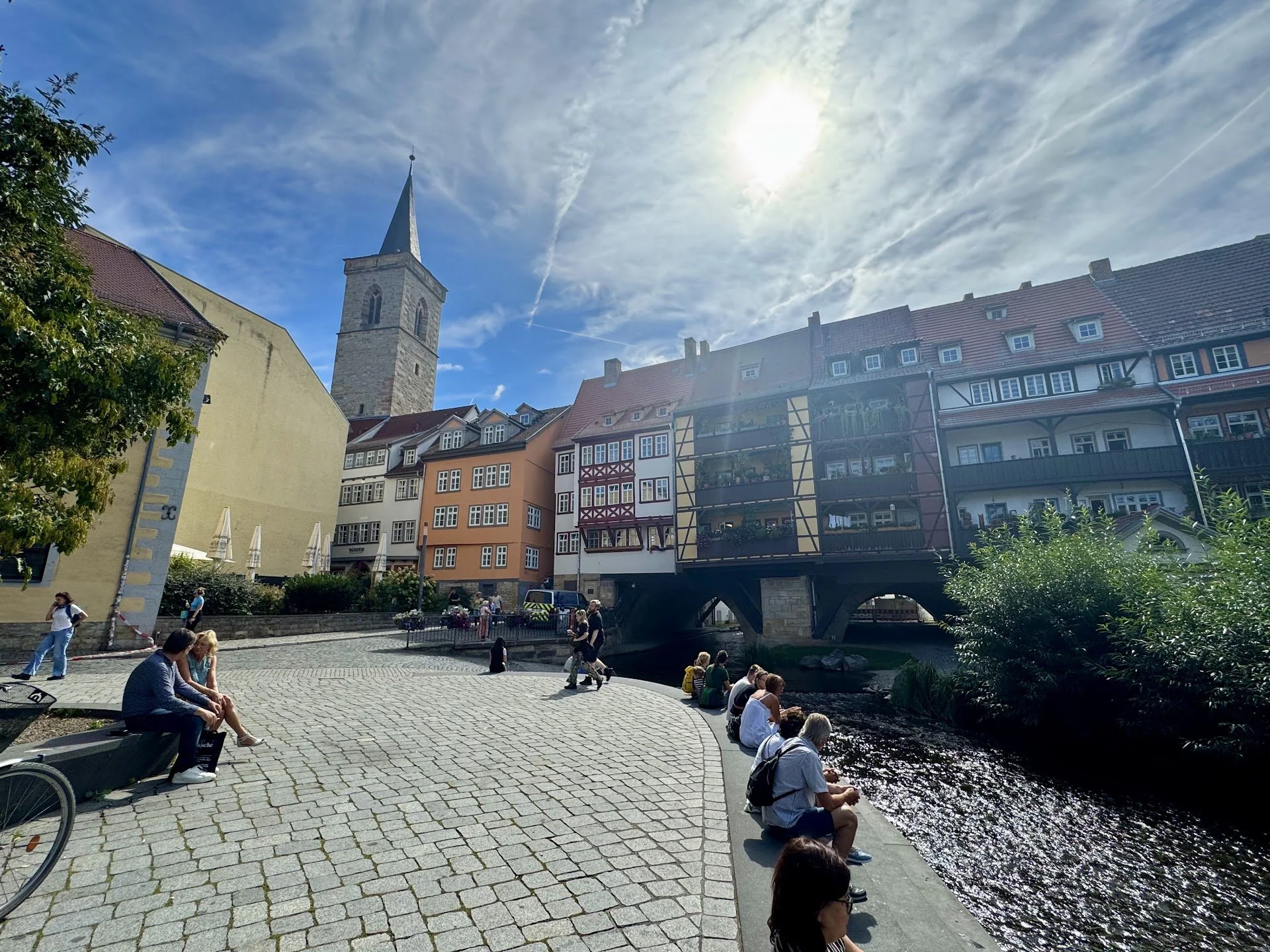 People sitting by the river in a European town with colorful buildings and a church steeple under a sunny sky.