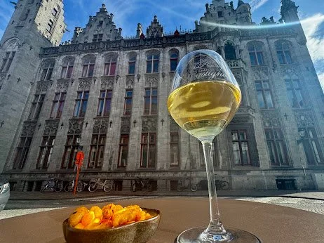 A glass of white wine and a bowl of cheese snacks on an outdoor table against a historic building.