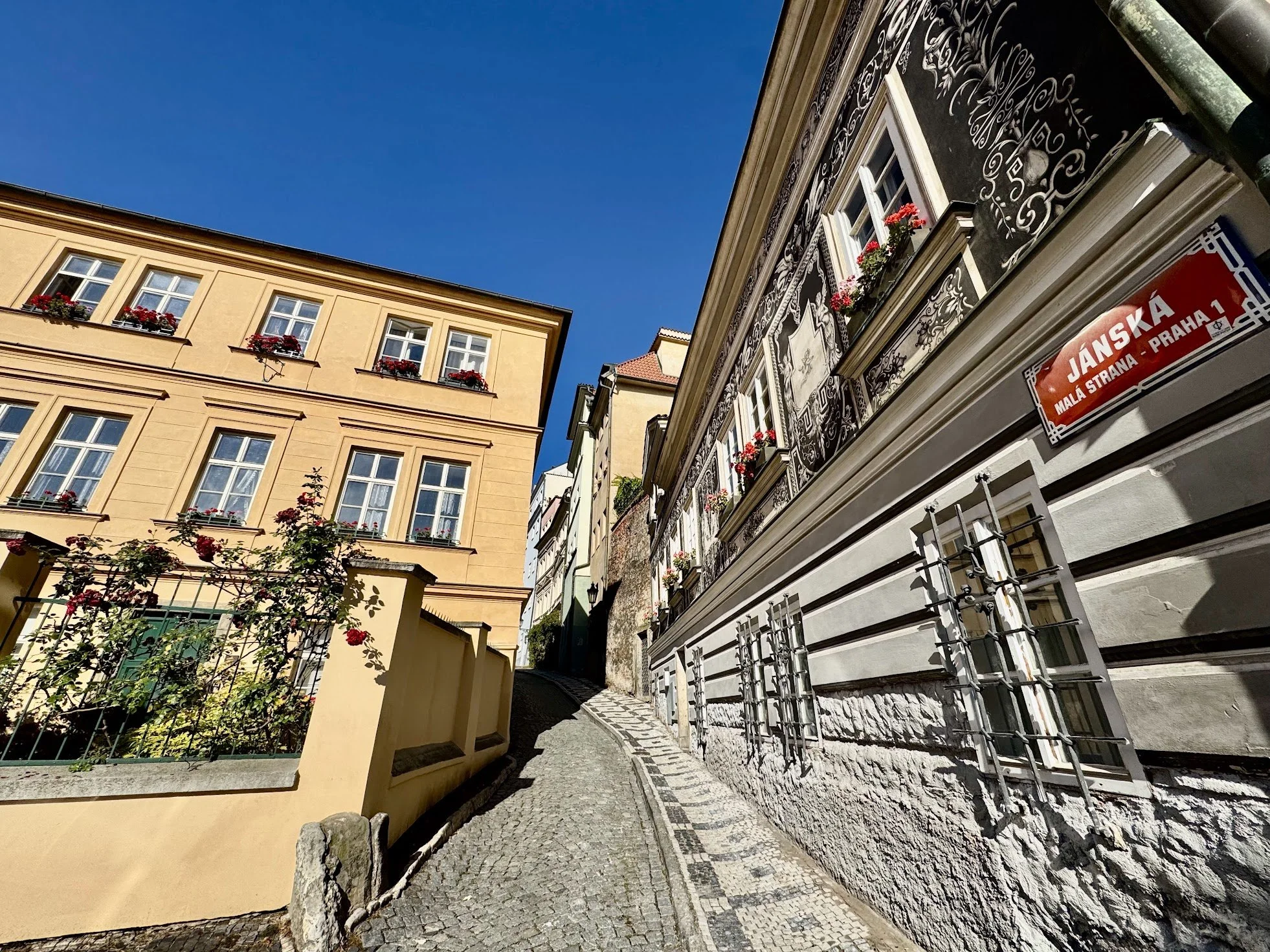 A narrow cobblestone street lined with buildings featuring flower boxes on the windowsills and ornate artwork on one building's facade, under a clear blue sky.