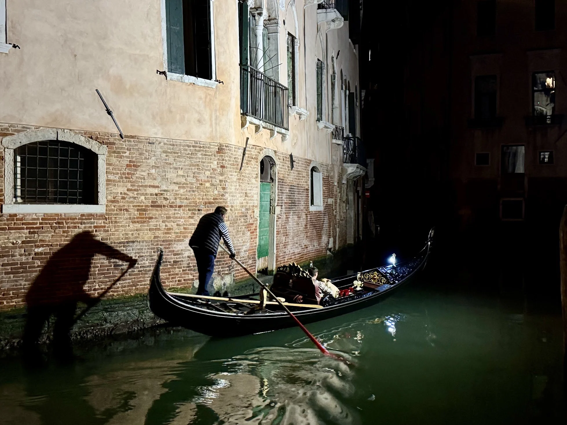 A gondola on a canal at night with a gondolier and two passengers, in Venice, Italy. The scene includes old brick and stucco buildings with windows along the canal.