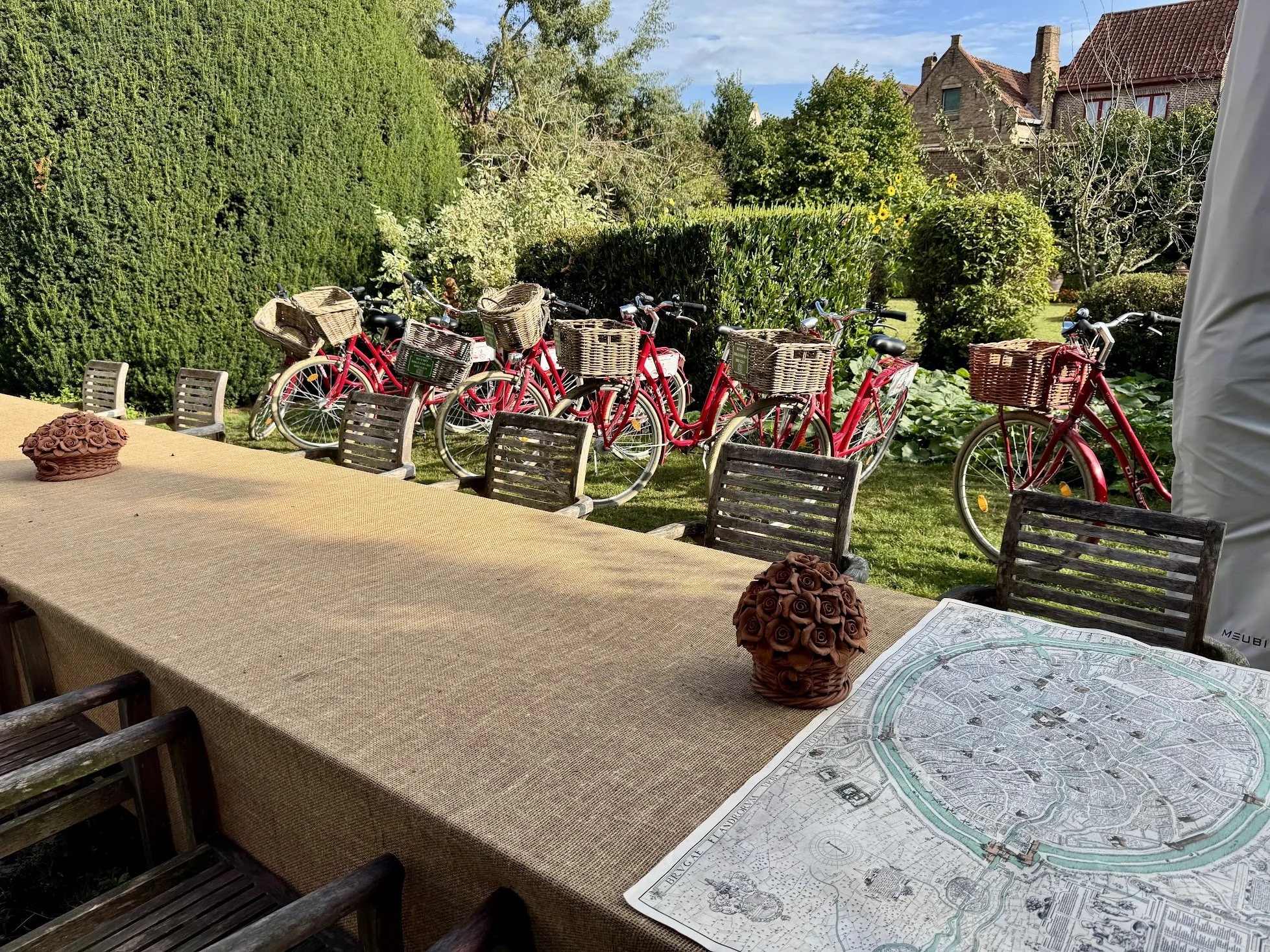 Outdoor table with chairs, decorative roses, and a large city map, overlooking a garden with several red bicycles parked near bushes and trees, in front of houses with brick walls and tiled roofs.