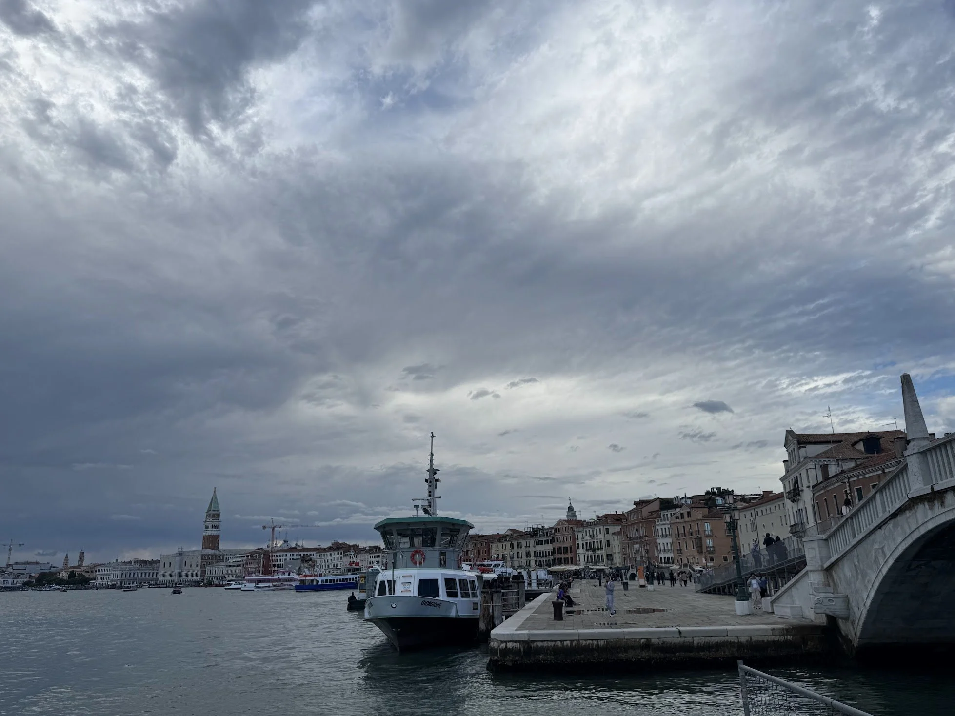 View of a harbor with boats docked along the pier, and historic buildings lining the waterfront under a cloudy sky.