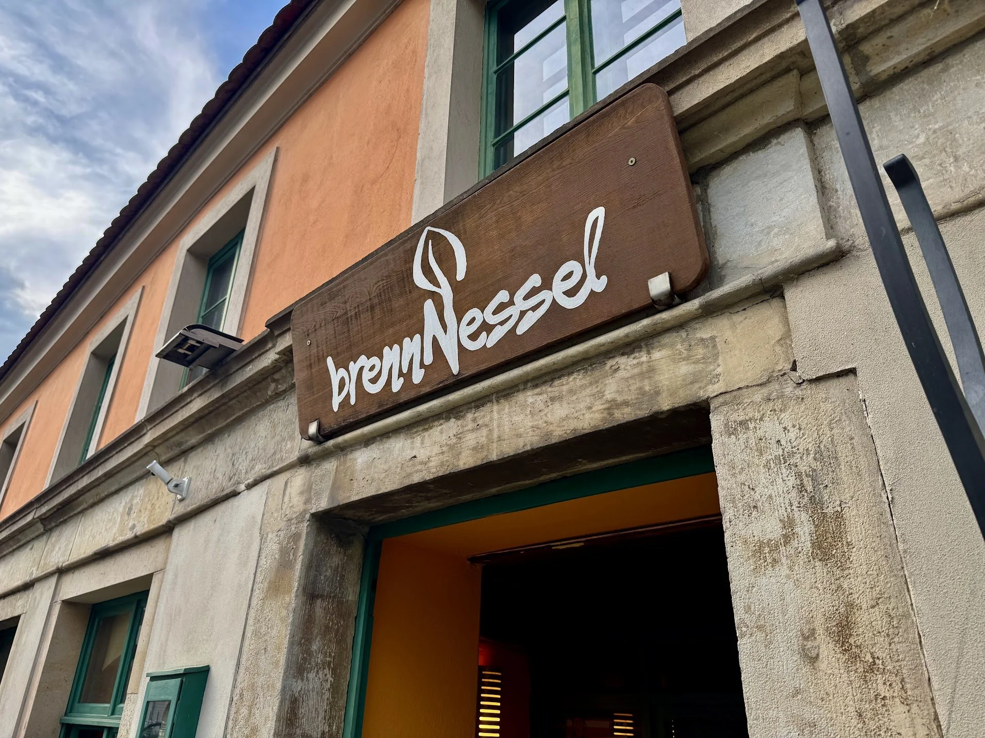 Exterior of a building with a wooden sign reading 'brennessel' above the entrance, green window frames, and a sunrise or sunset sky in the background.