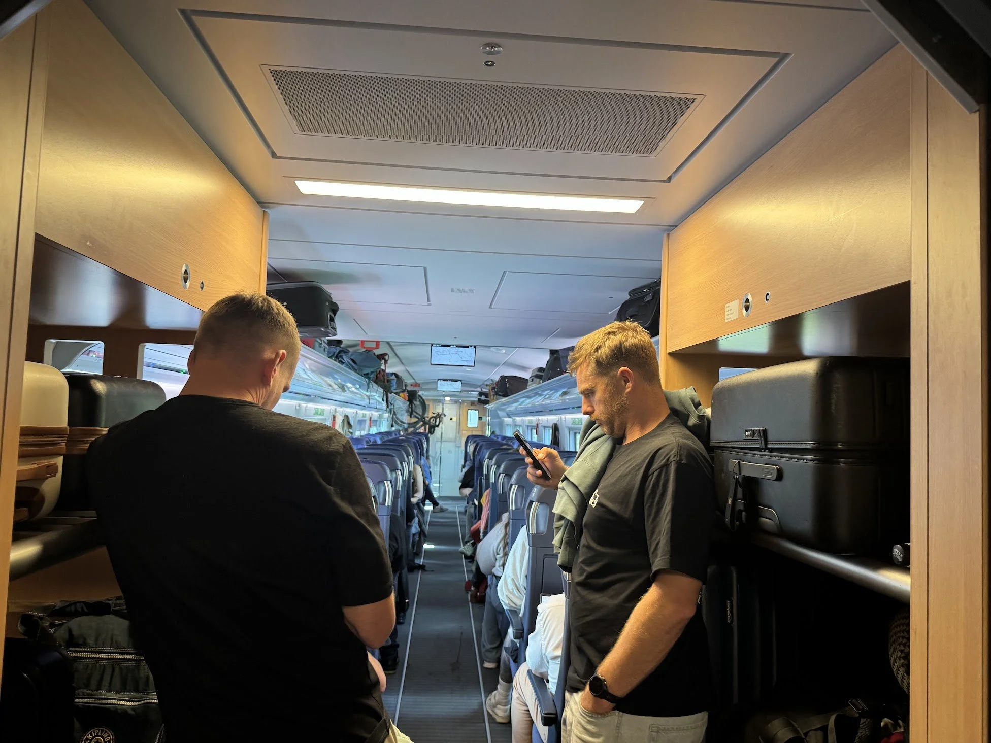 Interior of a train with passengers standing and sitting, using electronic devices, luggage on overhead racks, and seats arranged in rows.
