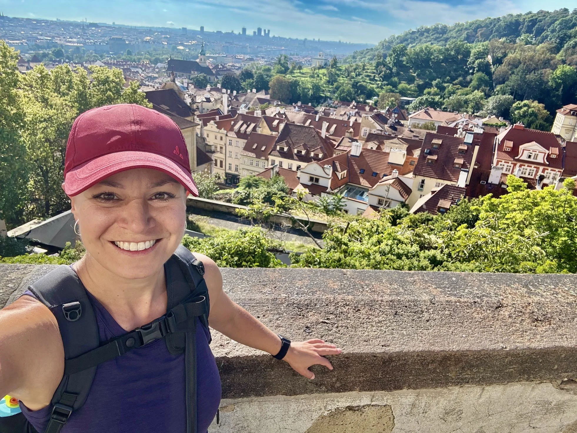 A woman smiling and taking a selfie with a scenic view of a European city with red-roofed buildings, green hills, and a church in the distance.