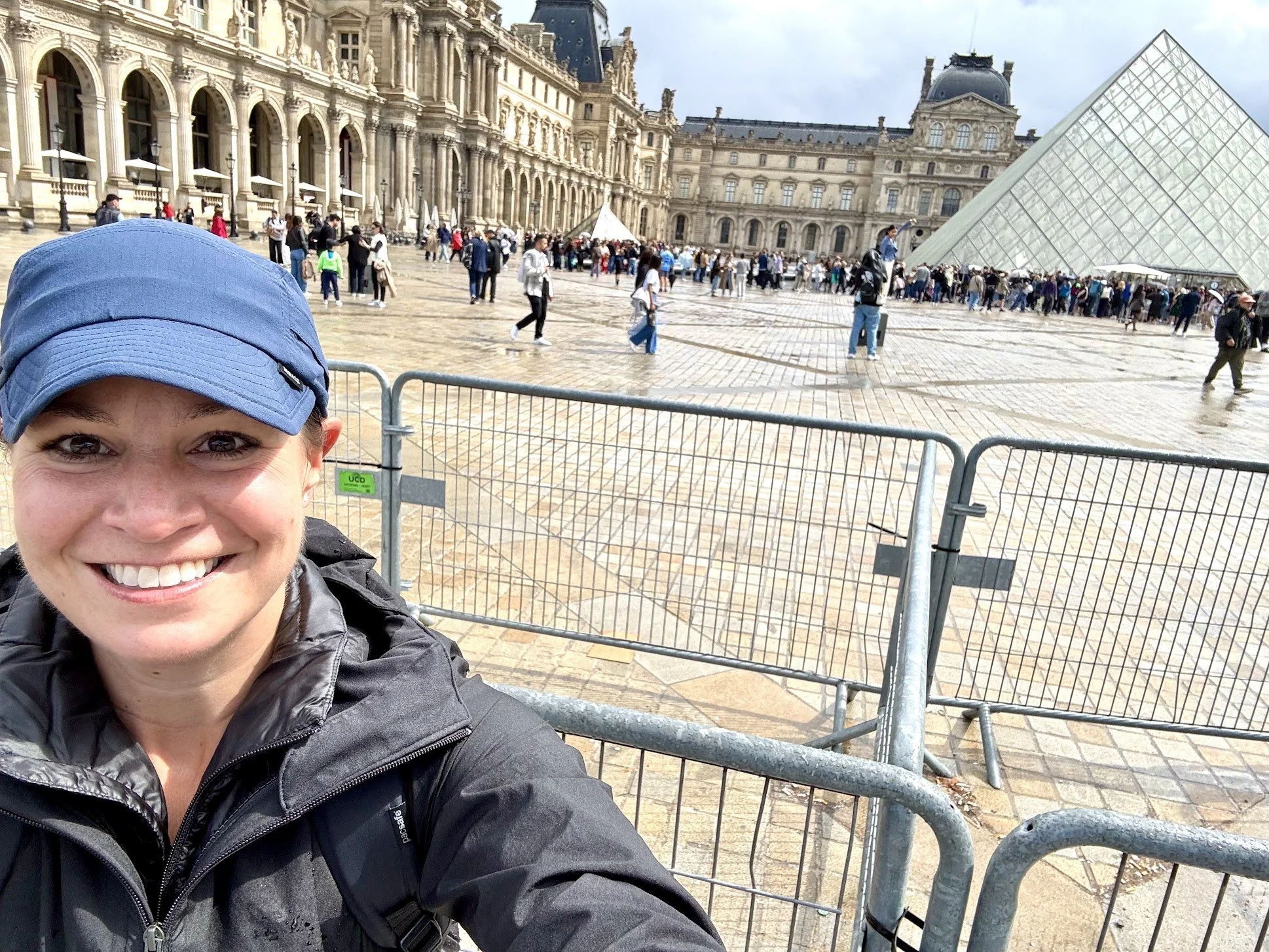 A woman smiling in front of a historic building with glass pyramid, crowd of people, and cloudy sky in the background.
