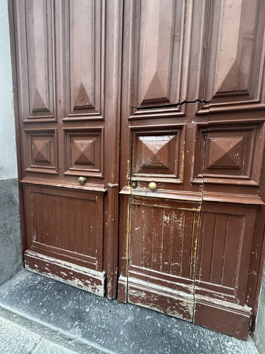 Old, weathered wooden double doors with ornate paneling, peeling brown paint, and a small brass knob on each door, set into a stone or concrete frame.