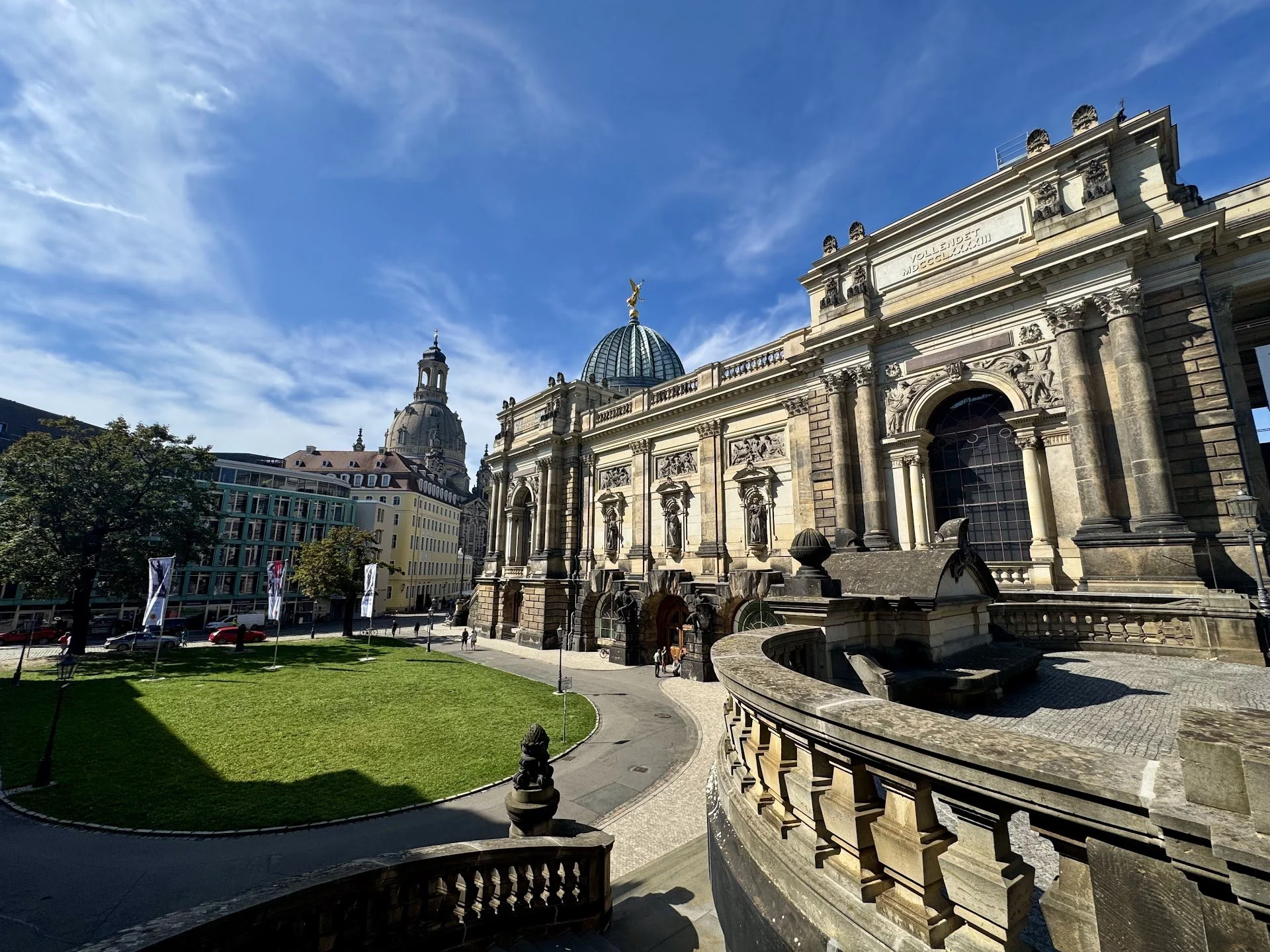 A historic building with ornate architectural details, columns, and domes, set against a bright blue sky with wispy clouds. A green lawn and a few people are visible in the foreground.