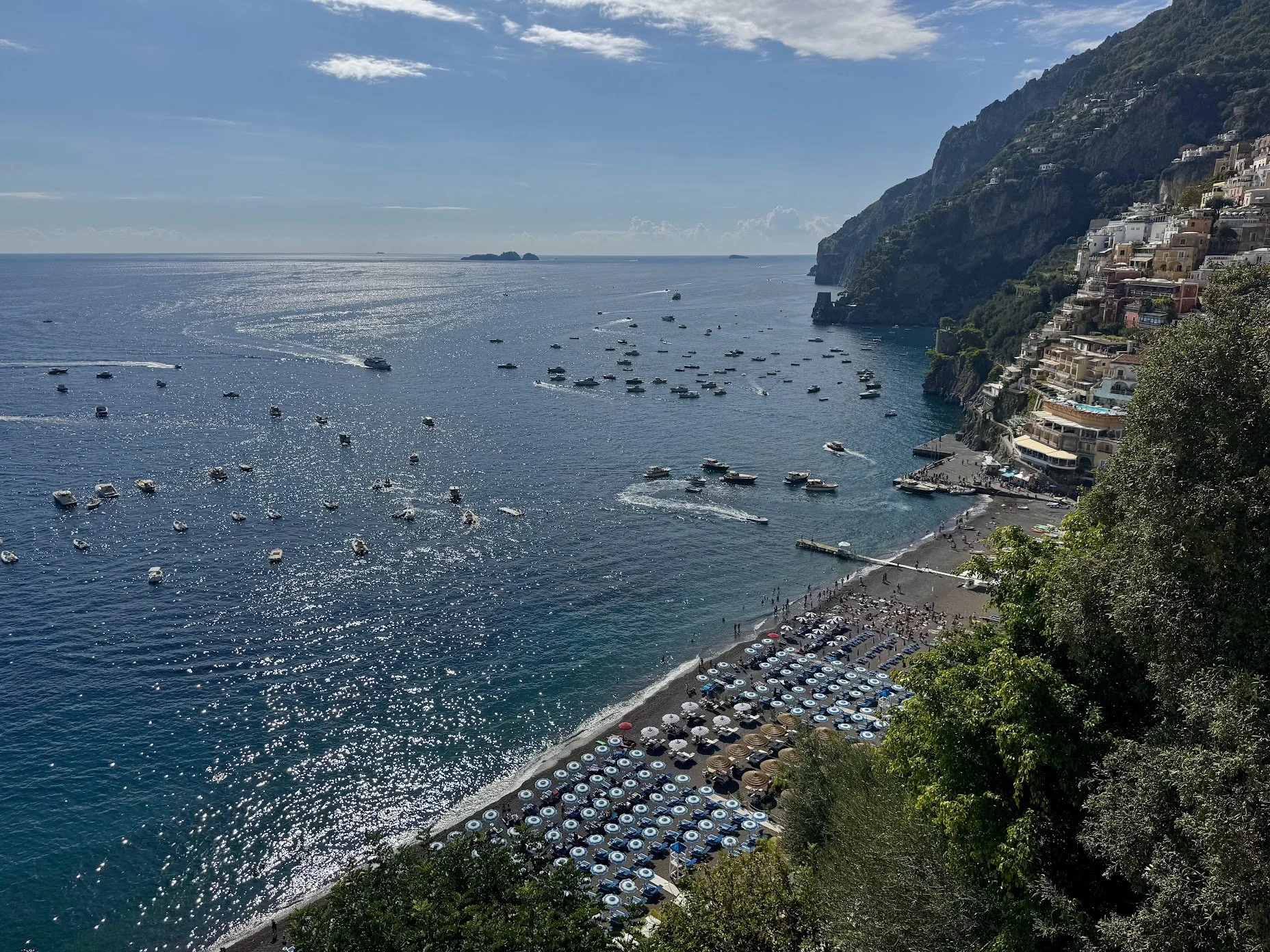 A coastal scene with a beach, boats on the water, and a hillside with houses.