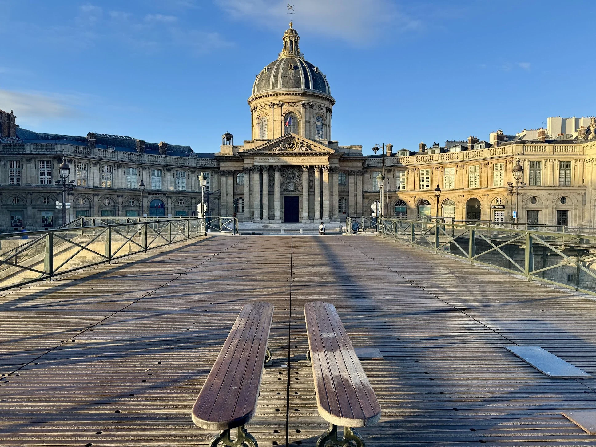 View of a historic building with a domed roof and classical architecture under a blue sky, taken from a wooden bridge with benches in the foreground.