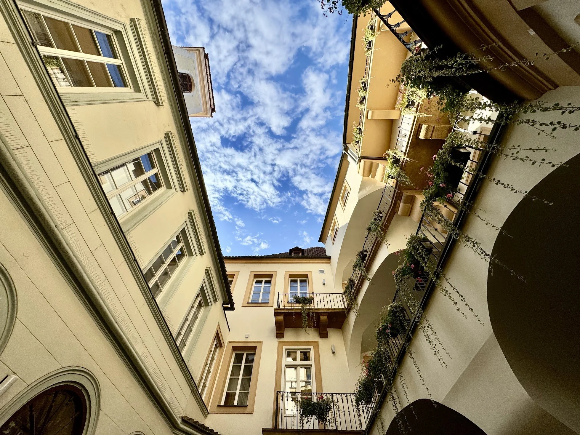An upward view of a historic European-style residential building courtyard with multiple windows, balconies with plants, and a blue sky with scattered clouds.