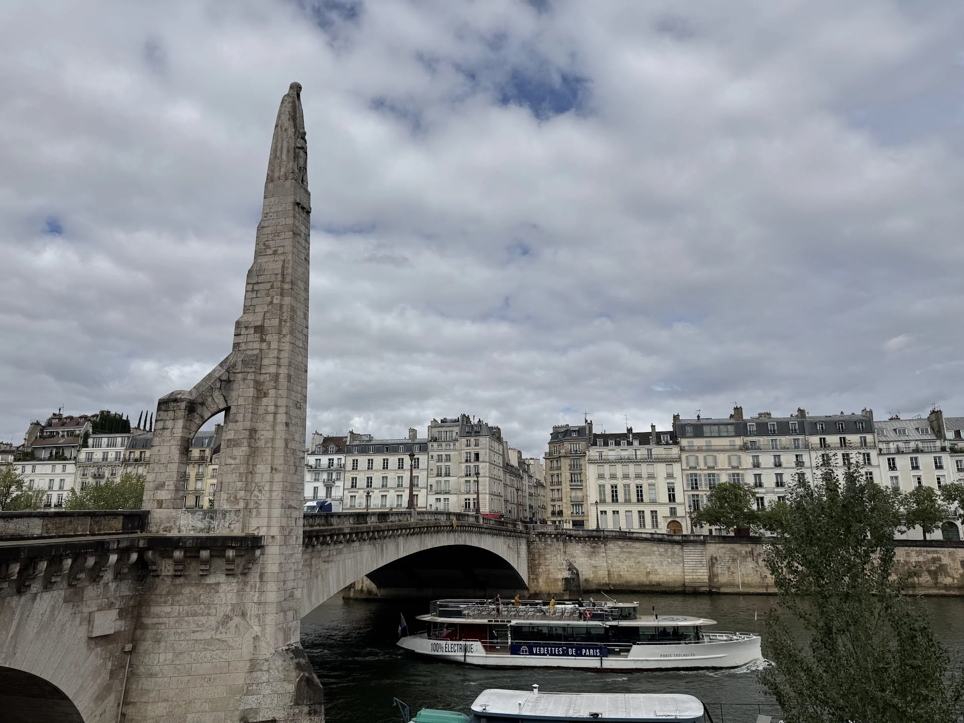 A Paris river scene with a stone bridge, a statue on the bridge, and a boat labeled 'VÉDETTES DE PARIS' on the water. Multi-story buildings line the riverbank under a partly cloudy sky.