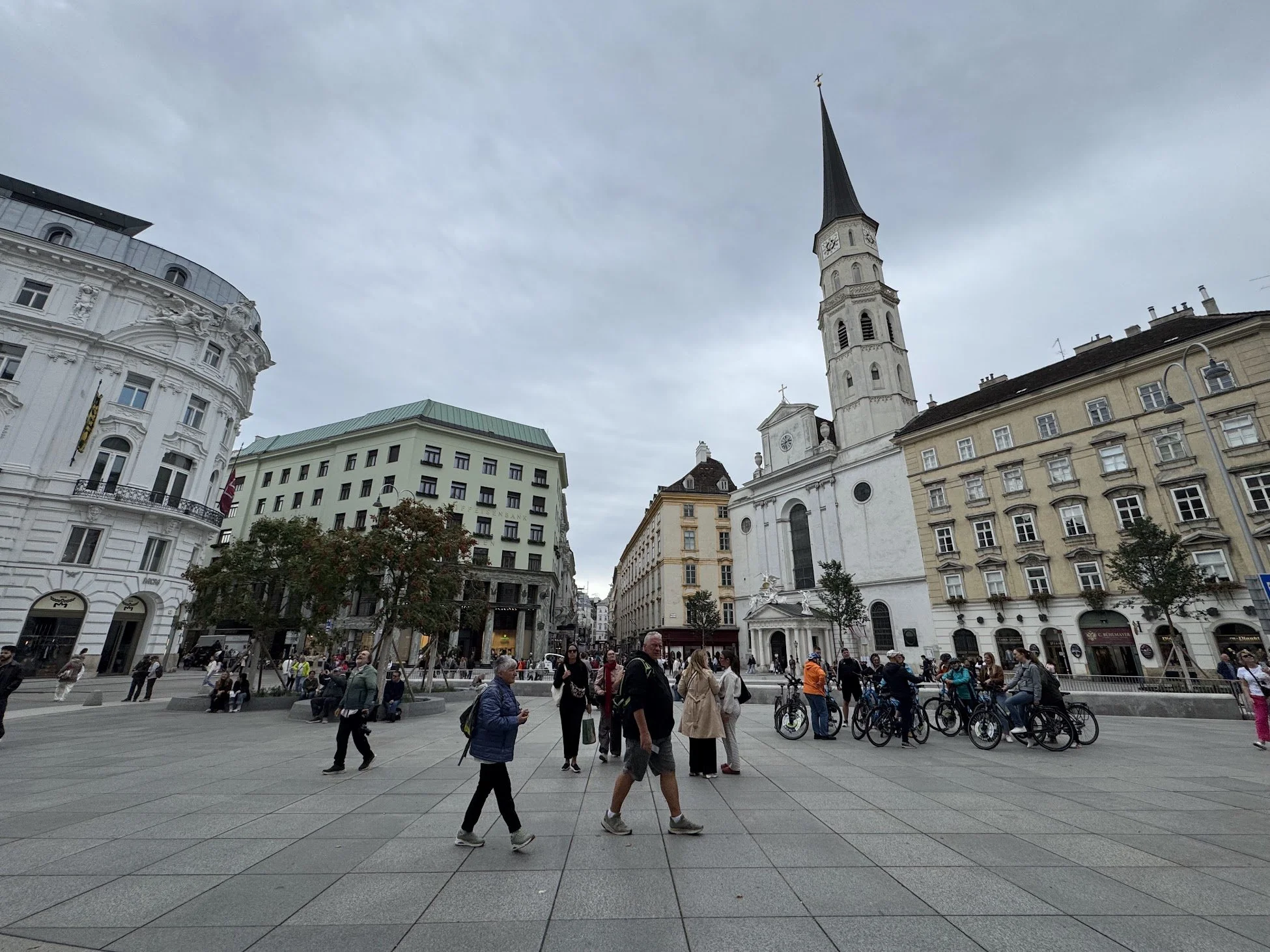 A busy European city square filled with pedestrians, some walking and others standing. Multiple bicycles are parked along the edge. Surrounding the square are historic and modern buildings, including a notable white church with a tall spire and a clo