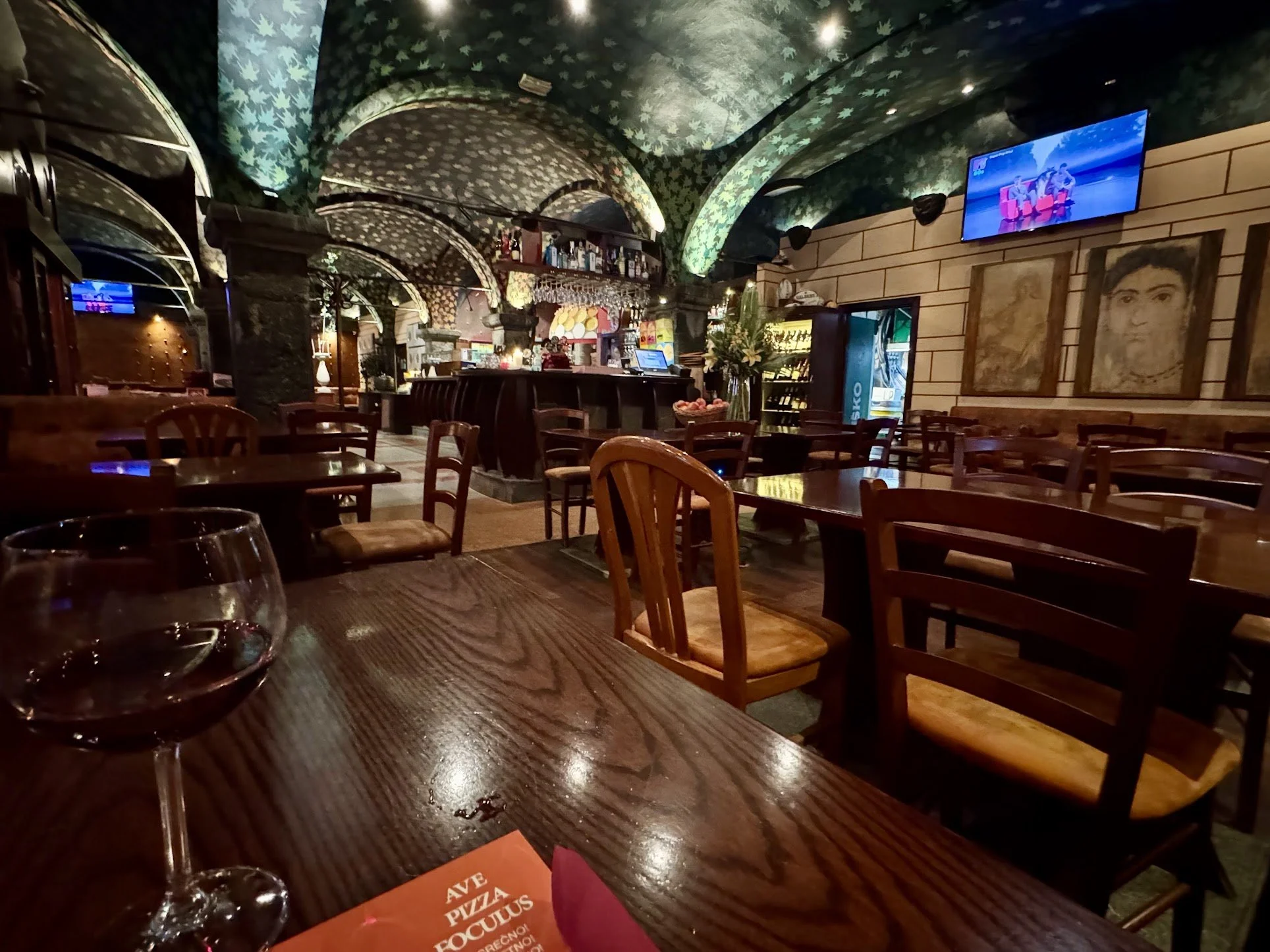 Interior of a restaurant or bar with wooden tables and chairs, a bar counter with bottles, and two televisions mounted on the wall showing a group of people on a boat. The ceiling has a green leafy pattern, and the wall features framed artwork.