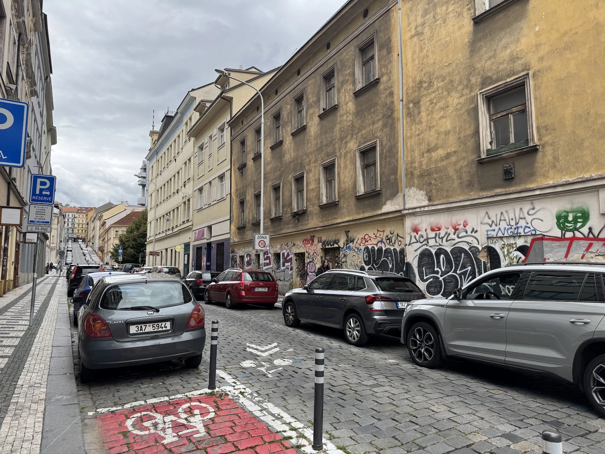 Street scene with multiple parked cars on cobblestone street, old buildings with graffiti, street signs including a reserved parking sign, a speed limit sign, and a oneway sign, under cloudy sky.