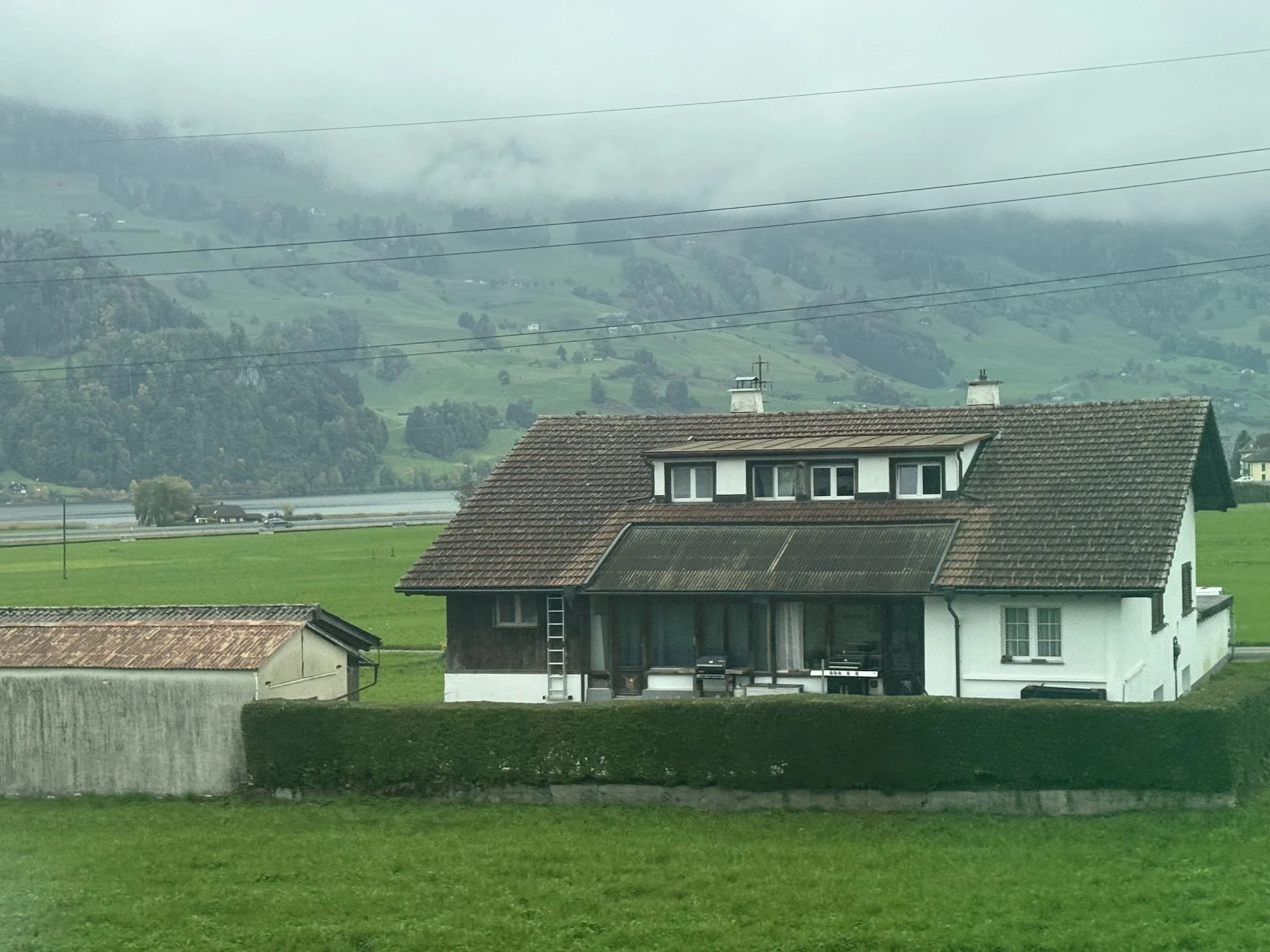 Green fields with a white house with a brown roof and two chimneys in a rural landscape with misty hills in background.