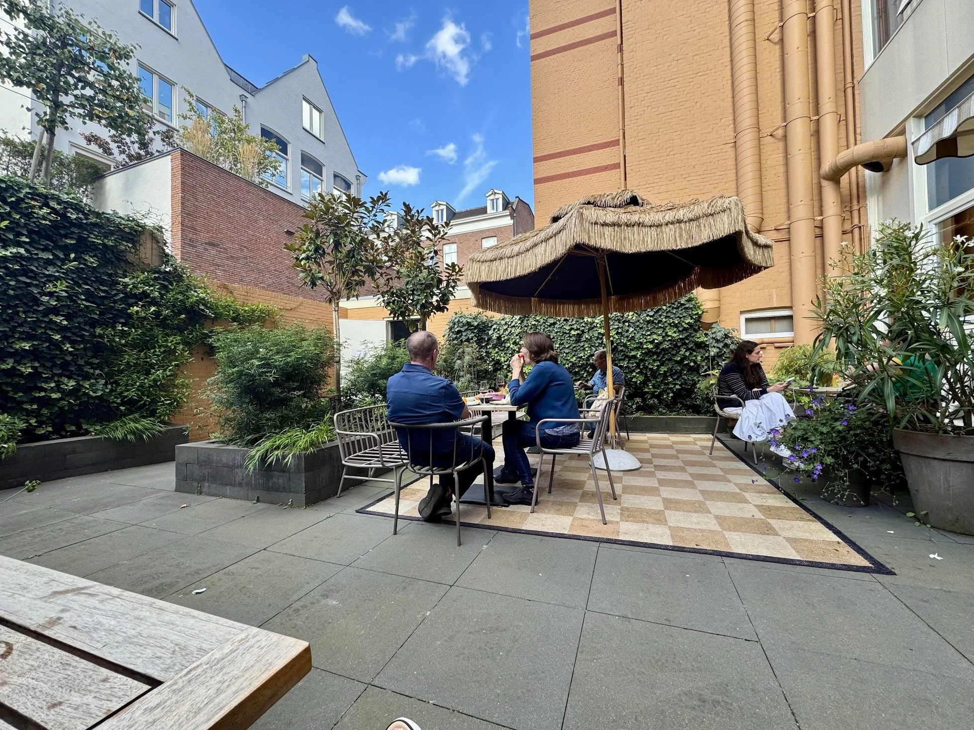 A small outdoor cafe garden with people sitting at tables under a straw shaded umbrella, surrounded by greenery and plants, with buildings and a blue sky in the background.