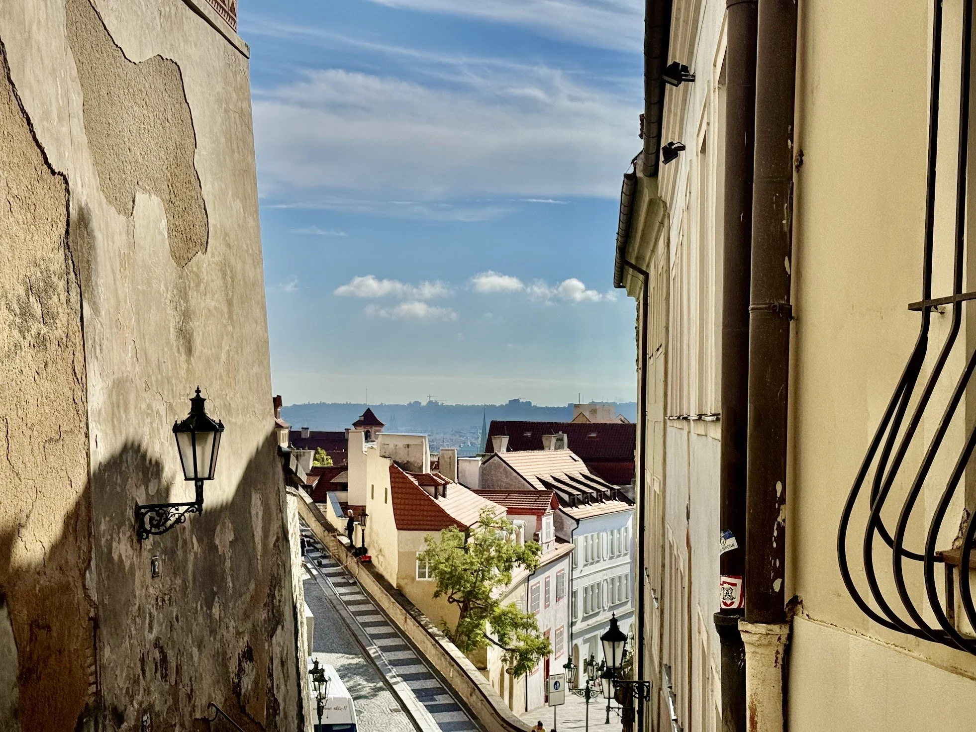 A view of a city street seen from a balcony or window, with historic buildings, street lamps, and a distant landscape with a water tower, under a partly cloudy sky.