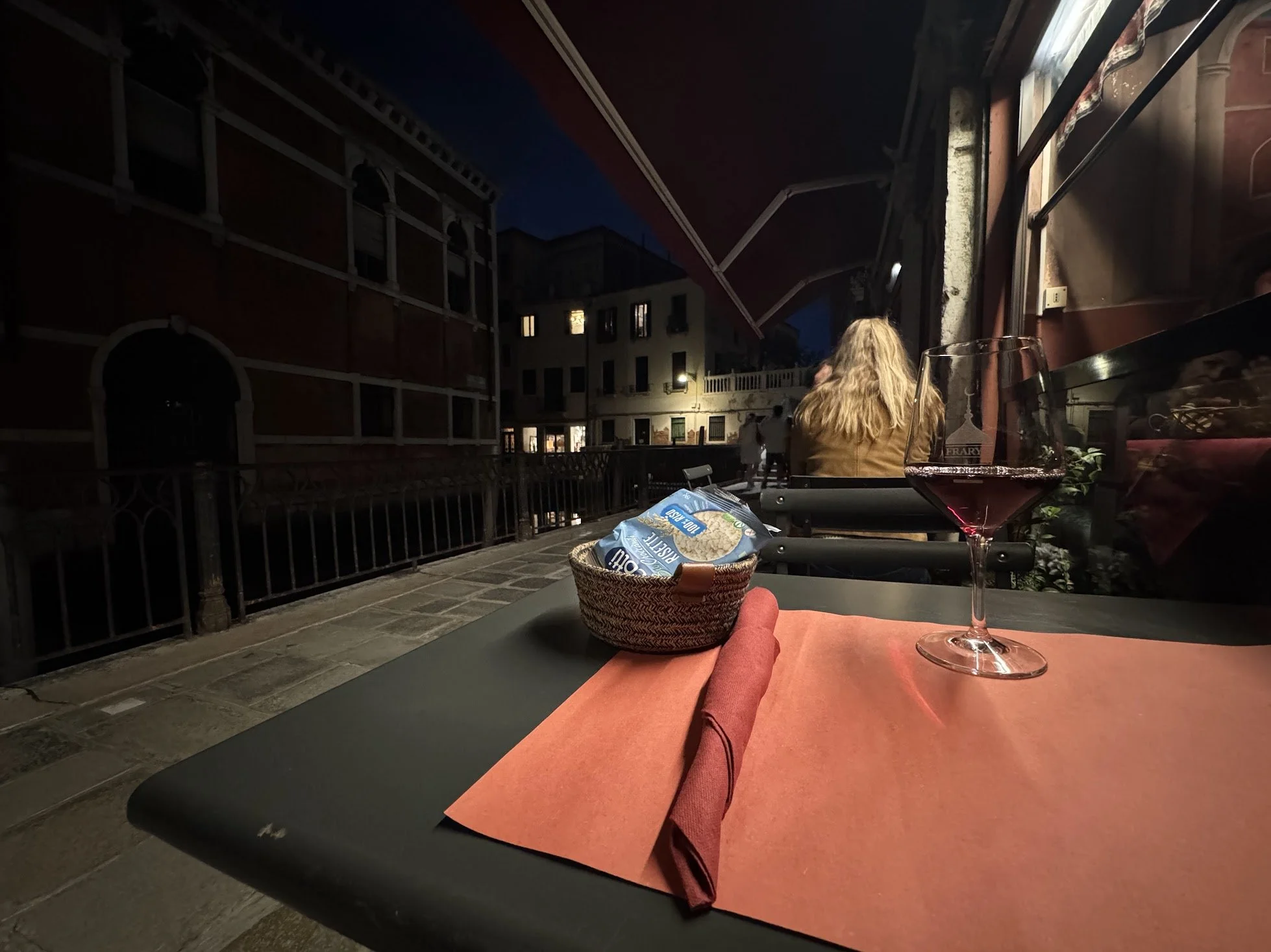 Nighttime scene at an outdoor dining area with a glass of red wine on a table, a basket of snacks, and a person's legs with red pants. In the background, a woman with blonde hair sits facing away, and other people are walking nearby on a balcony line