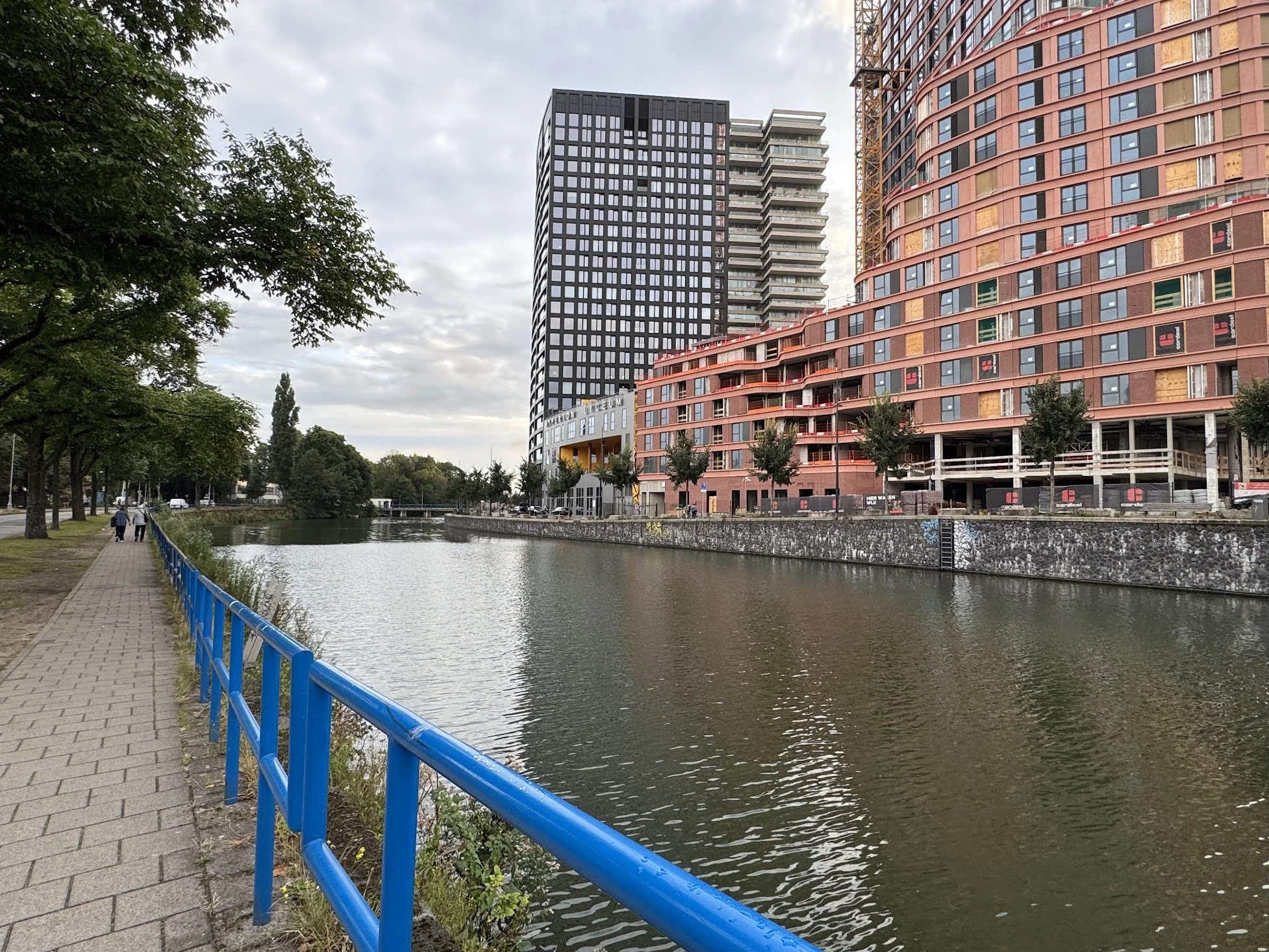 A cityscape with modern high-rise buildings along a river, with a paved walkway and blue railing on the left side, trees lining the riverbank, and overcast sky.