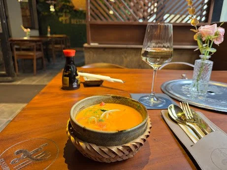 A bowl of soup, a glass of white wine, a small vase with pink flowers, soy sauce bottle, white chopsticks, and silverware on a wooden table in a restaurant setting.