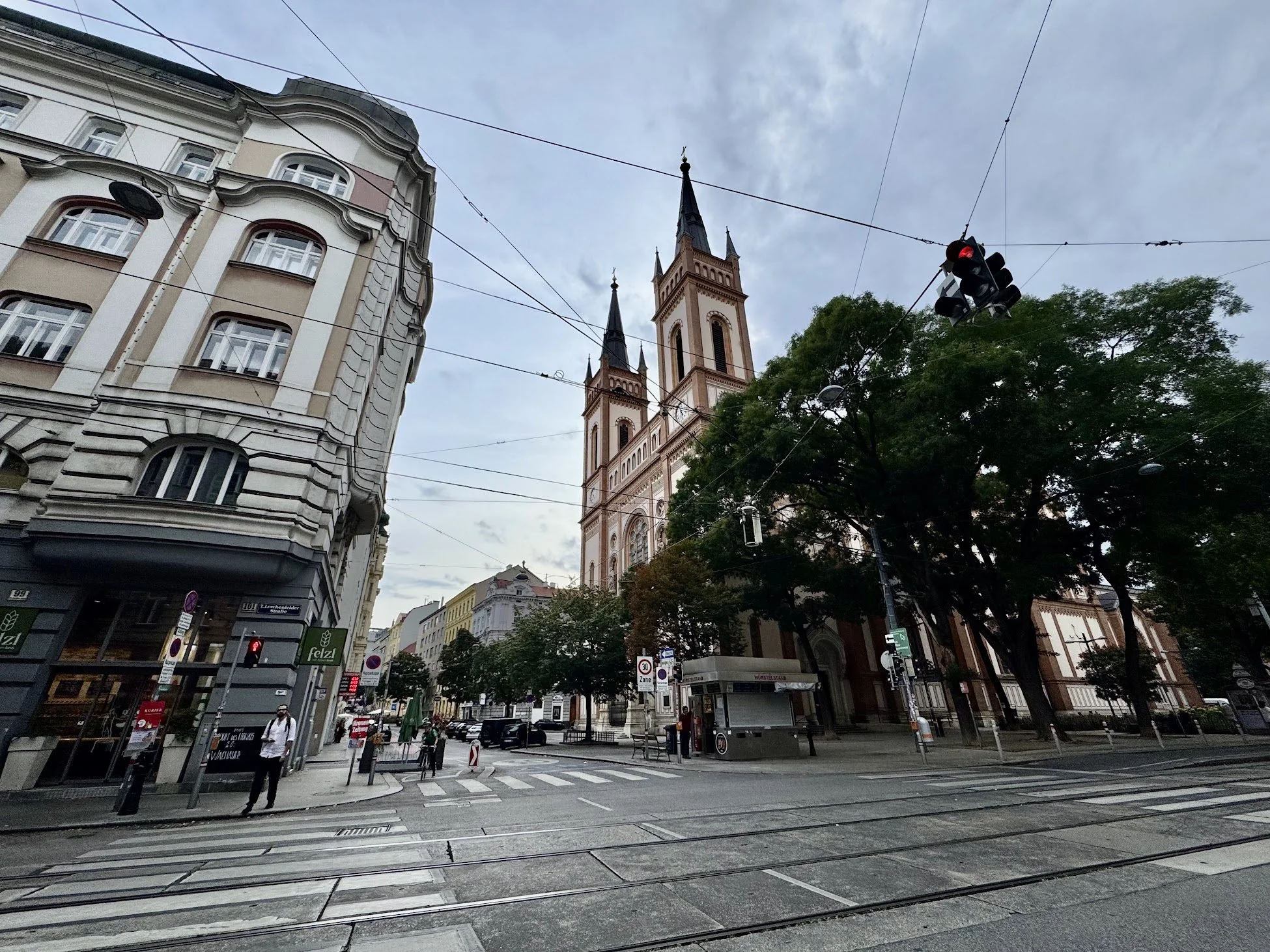 A city street scene with a historic church with tall spires in the background, traffic lights, streetcars, cars, pedestrians, and buildings under a cloudy sky.