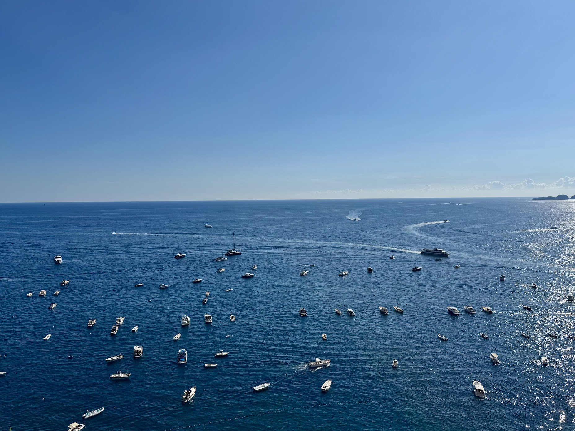 Aerial view of a large body of water with numerous boats, yachts, and speedboats scattered across the surface, under a clear blue sky with sunlight reflecting off the water.