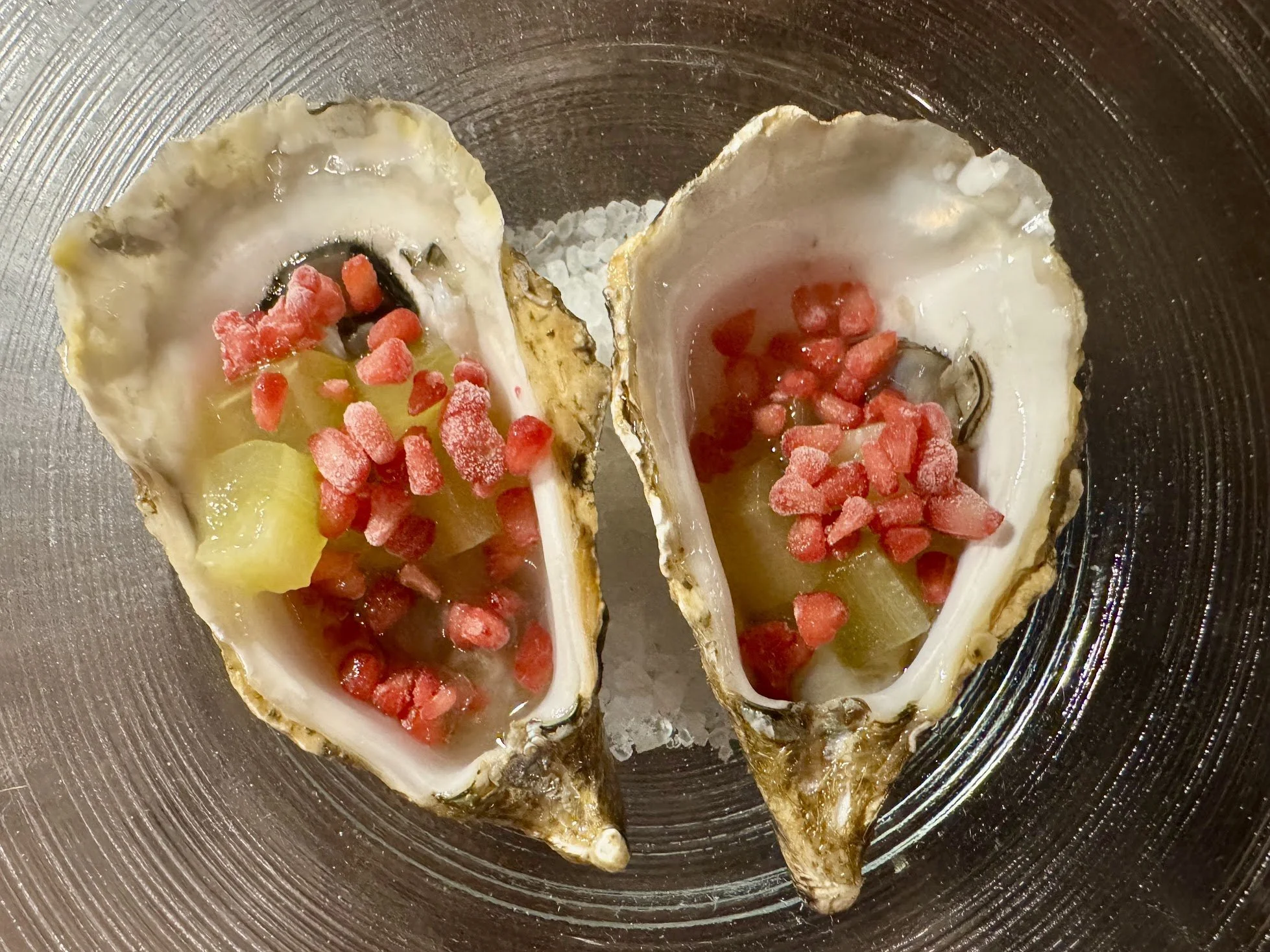 Open oyster shells filled with lemon juice, pink fish roe, and a spoon for eating, placed on a textured metal plate.