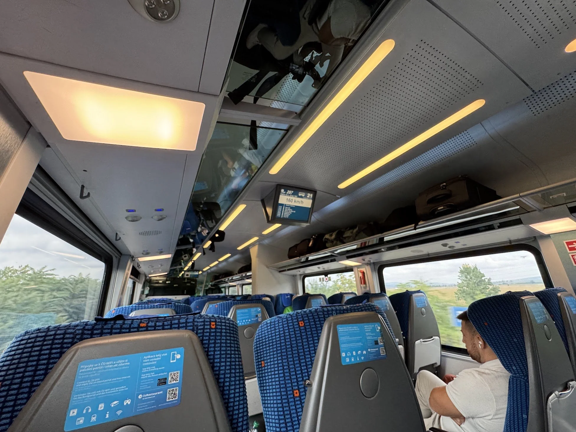 Interior of a train carriage with blue patterned seats, digital screens, overhead compartments, and passengers seated and standing, viewed from the aisle.