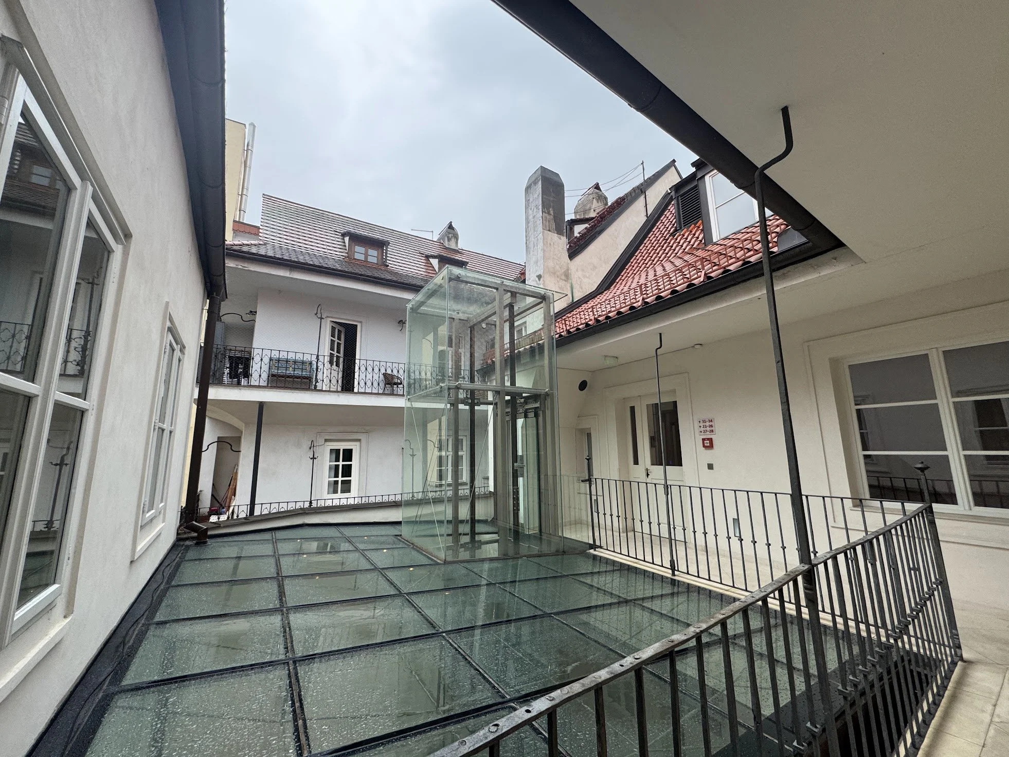 A view of an indoor courtyard with glass flooring, surrounded by white walls and black metal railings, and featuring an elevator shaft made of glass. The courtyard is enclosed by buildings with white walls and red-tiled roofs, with a cloudy sky overh