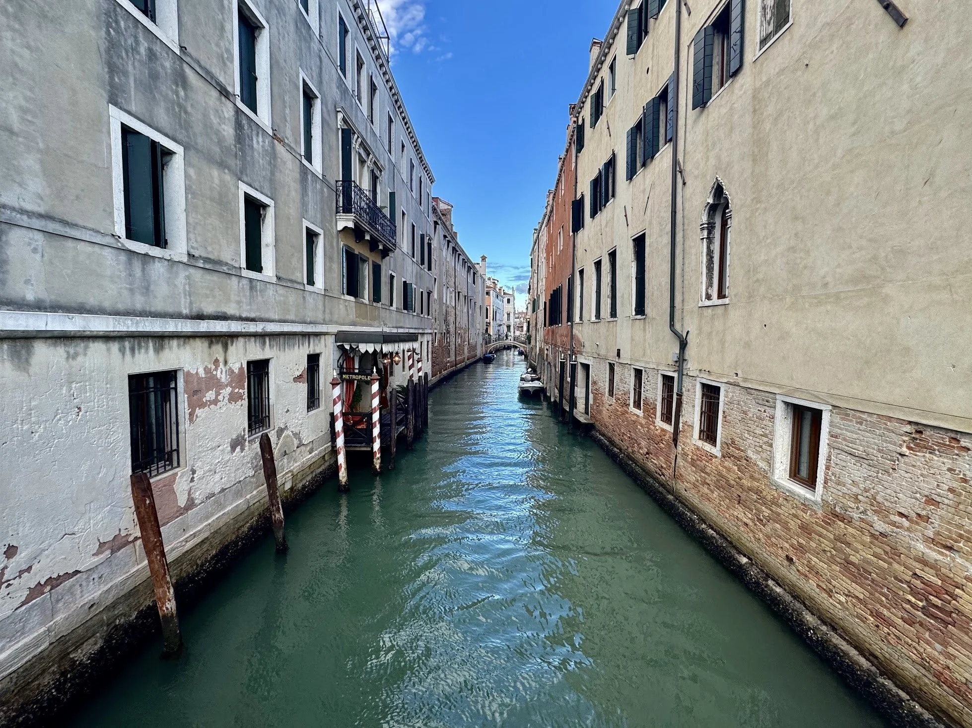 A narrow canal in Venice, Italy, surrounded by old buildings with weathered facades and small windows. The water is green and calm, with boats docked along the edges under a bright blue sky.