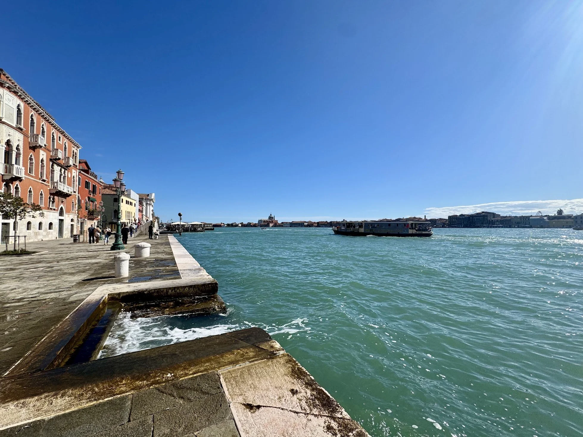 A waterfront promenade with colorful historic buildings, a boat on the water, and a clear blue sky.