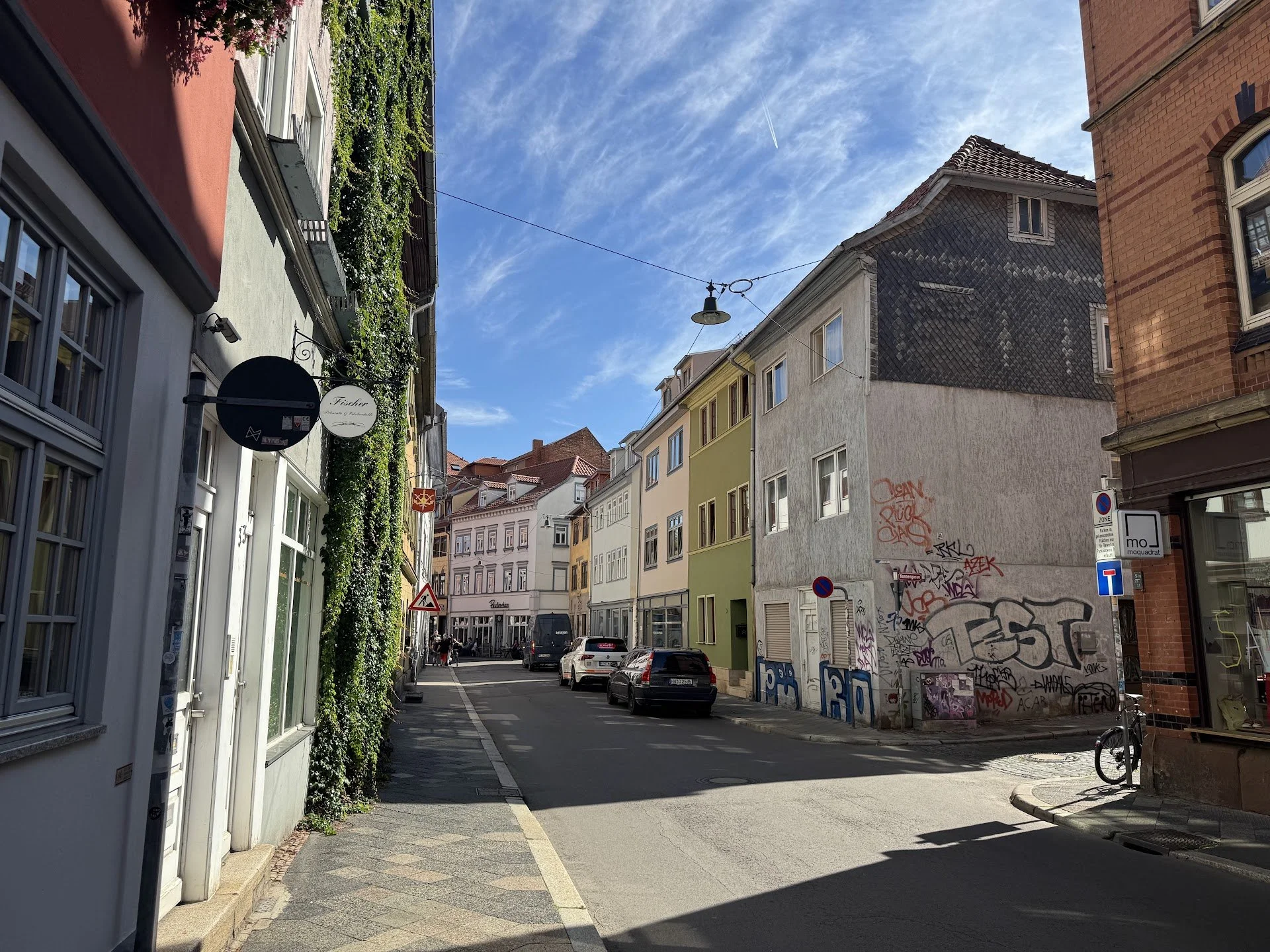 A quiet urban street scene with colorful multi-story buildings, parked cars, and graffiti on a wall, under a blue sky with wispy clouds.