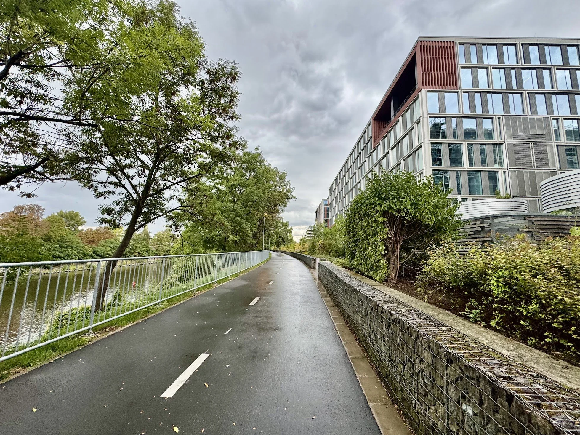 A paved cycling or walking path bordered by green bushes and trees on the left and right, with a modern multi-story building with glass windows on the right, under a cloudy sky.