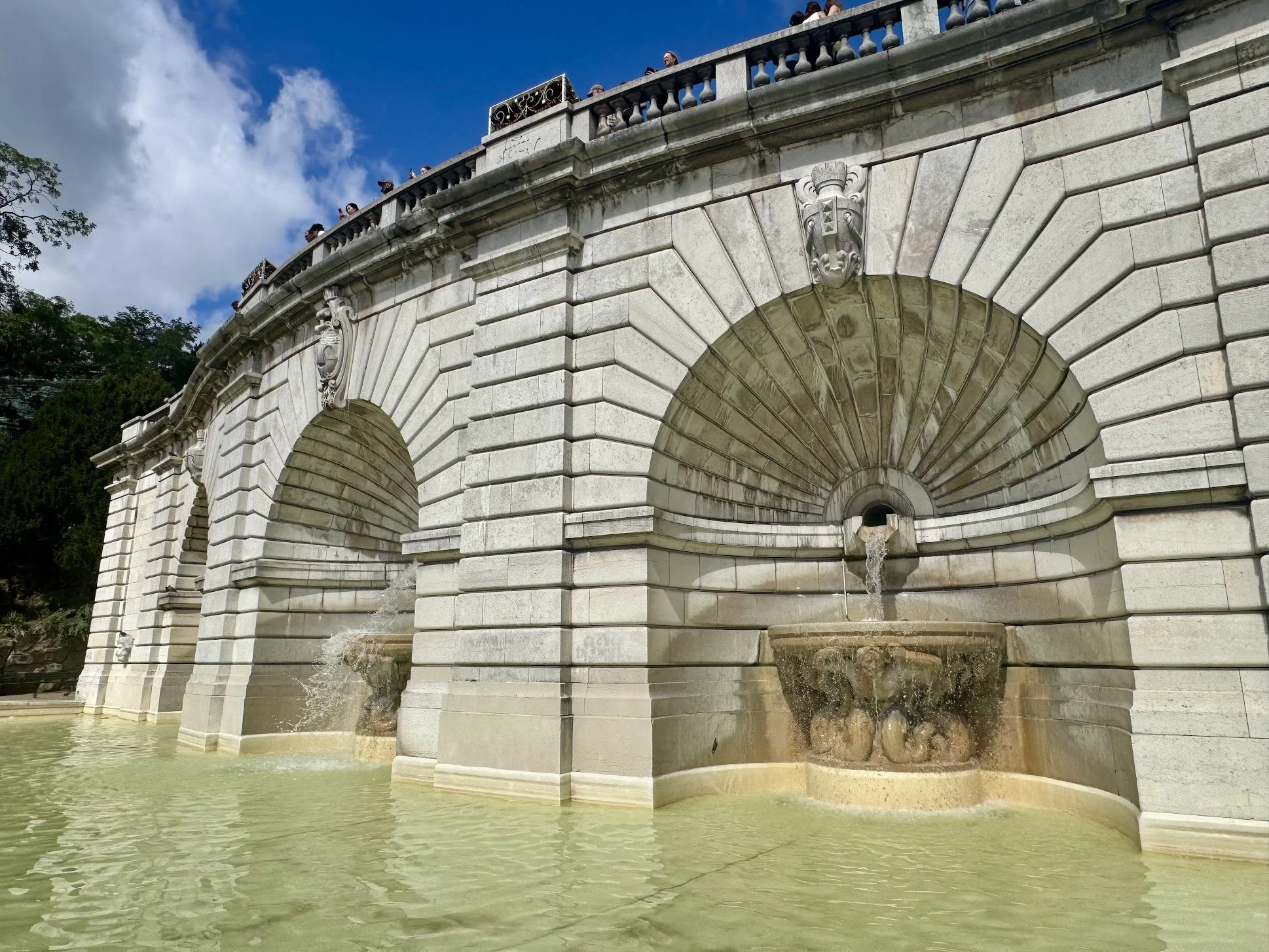 A historic stone fountain with arches and sculpted heads, water flowing from the mouth of the sculptures into a greenish pool, under a partly cloudy blue sky.