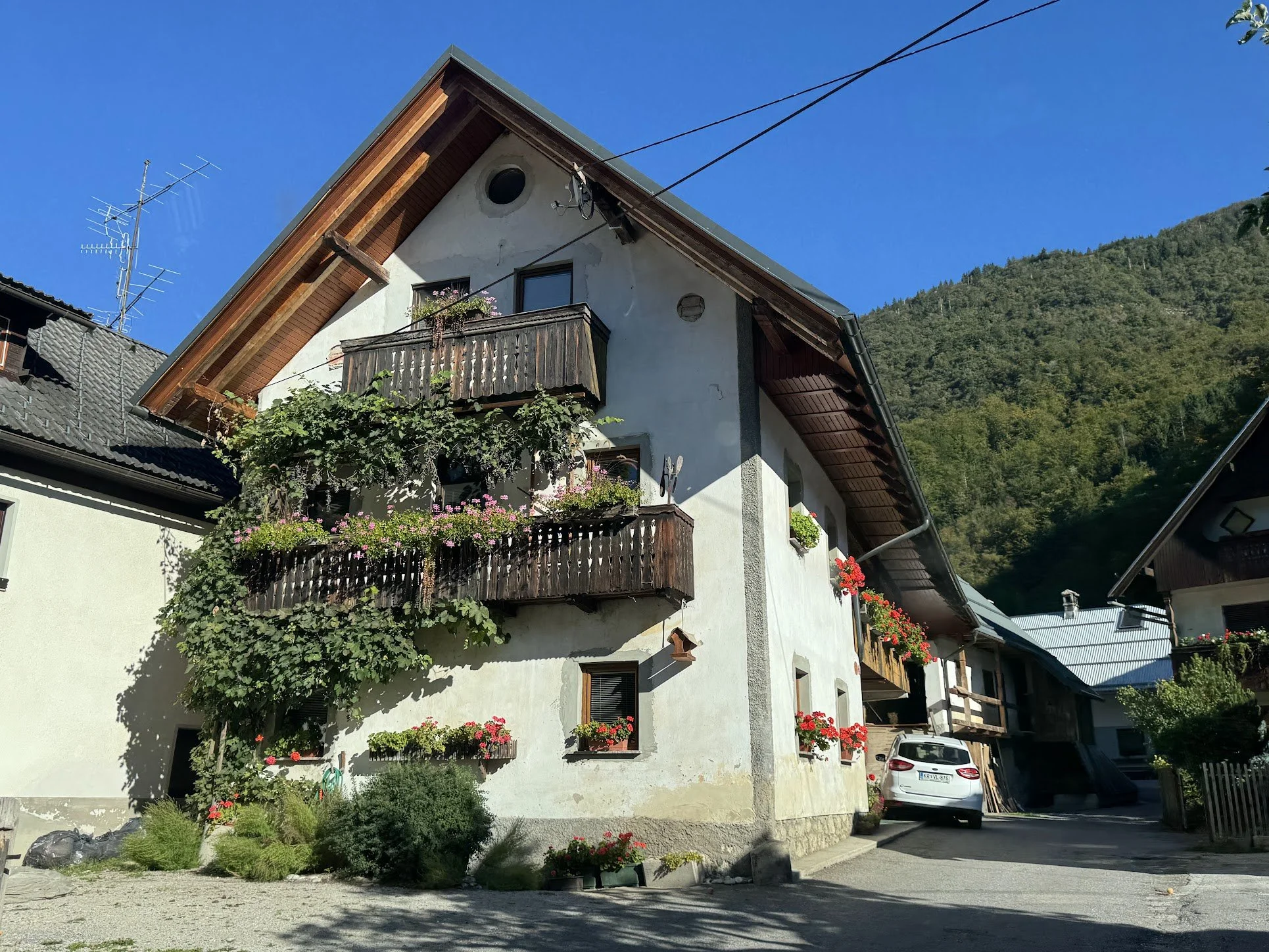 A residential house with white walls and a sloped roof, decorated with flower boxes on the balconies, greenery at the base, in a mountainous area under a clear blue sky.