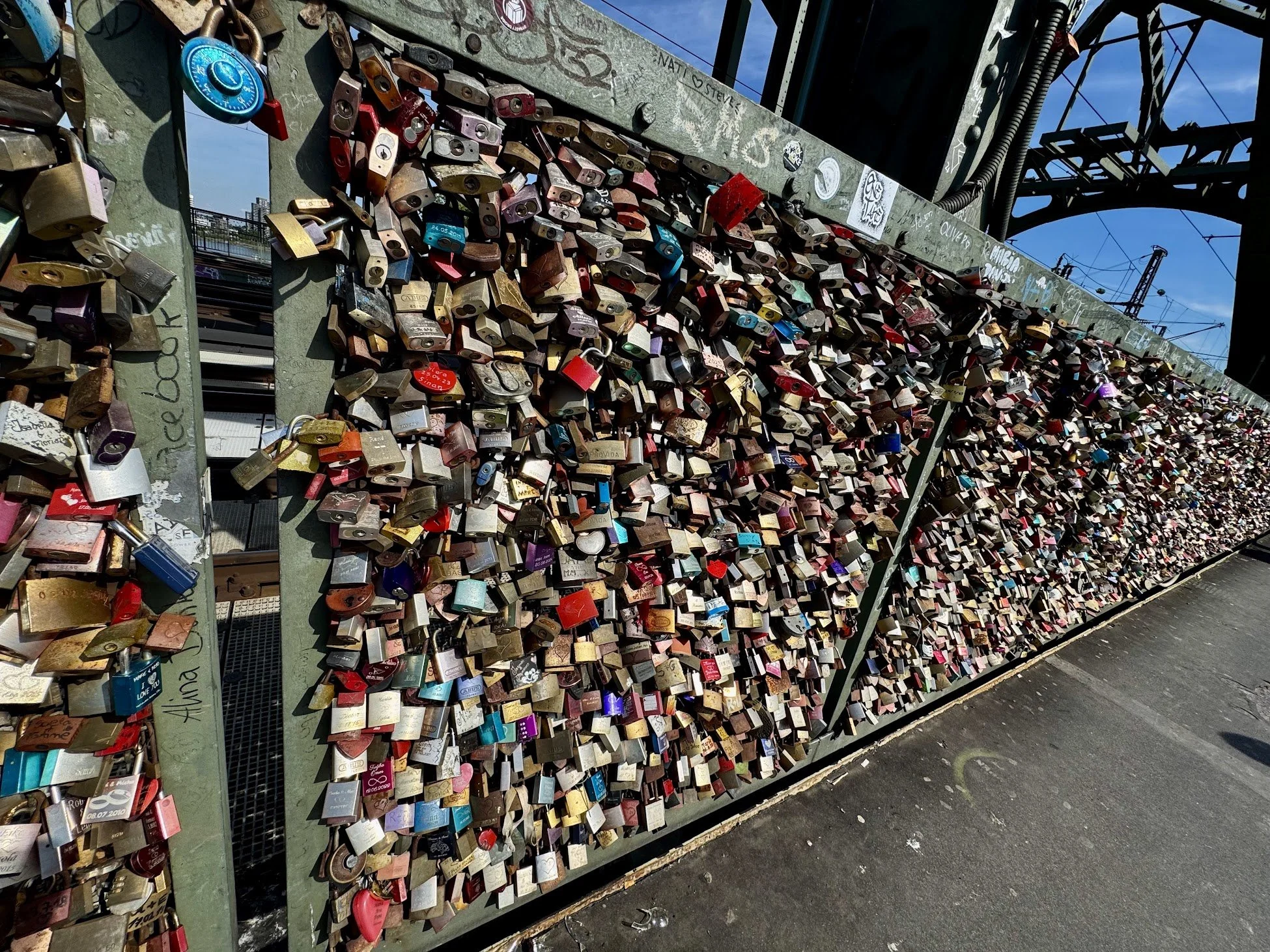 Wall covered with numerous colorful love locks attached to a metal structure under a bridge, with graffiti and electrical wires in the background.