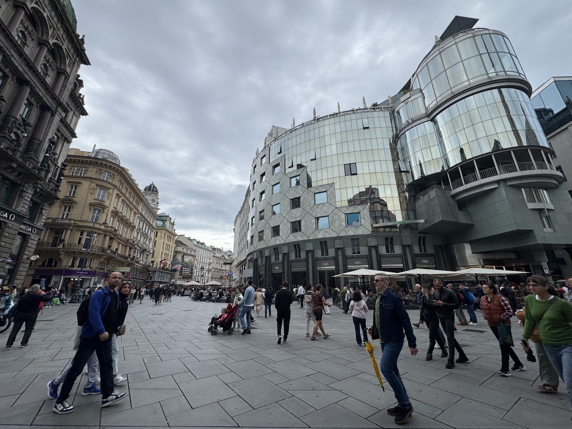 A busy city square filled with people walking, shopping, and socializing, surrounded by historic and modern buildings under a cloudy sky.