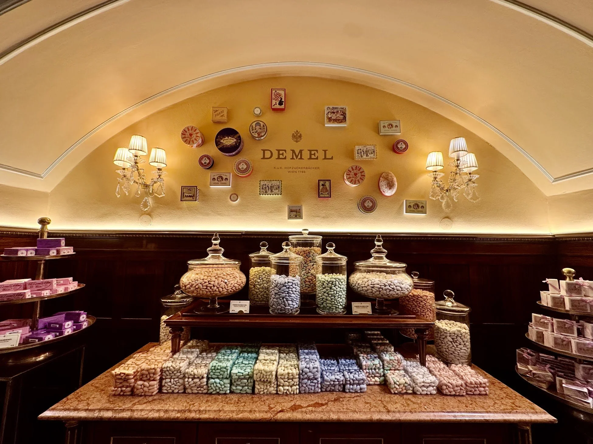 Display of colorful candies in glass jars at a bakery or candy shop, with labeled containers and decorative wall behind.