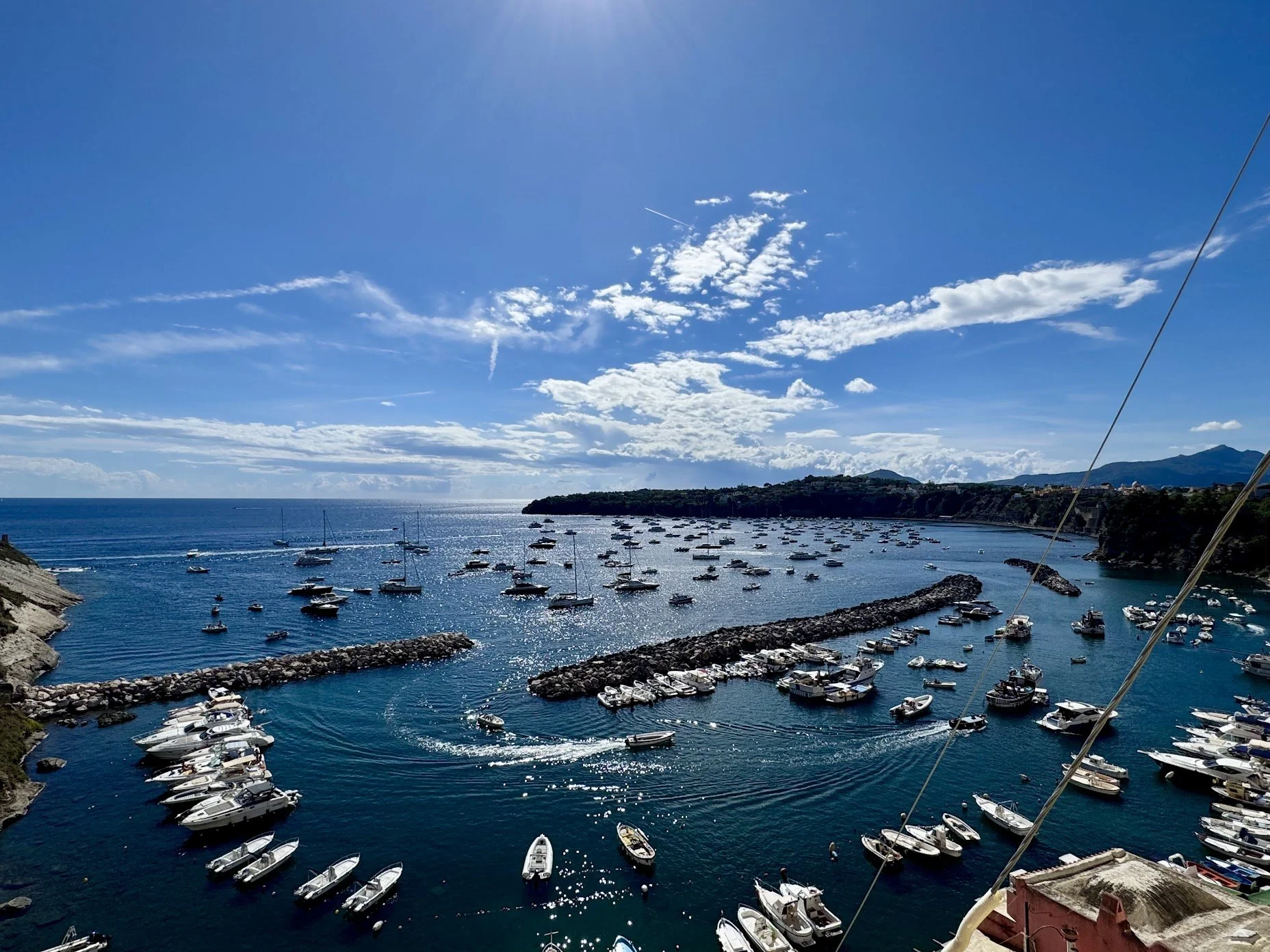 A harbor filled with numerous boats and yachts, surrounded by rocky breakwaters, with a small island or peninsula in the background under a blue sky with scattered clouds.