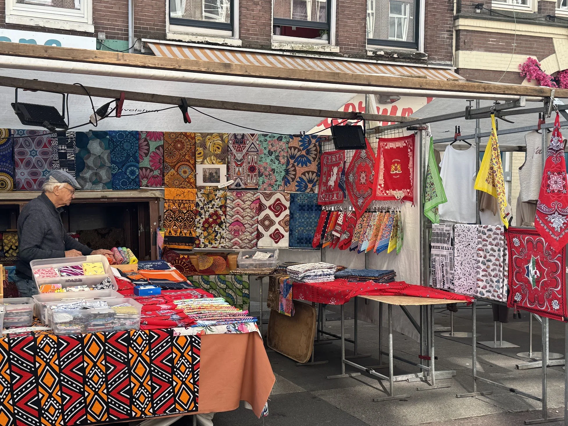 A street market stall displays colorful patterned textiles, clothing, and accessories. A man stands at the left, sorting items. Behind the stall, there is a brick building with windows and a striped awning.