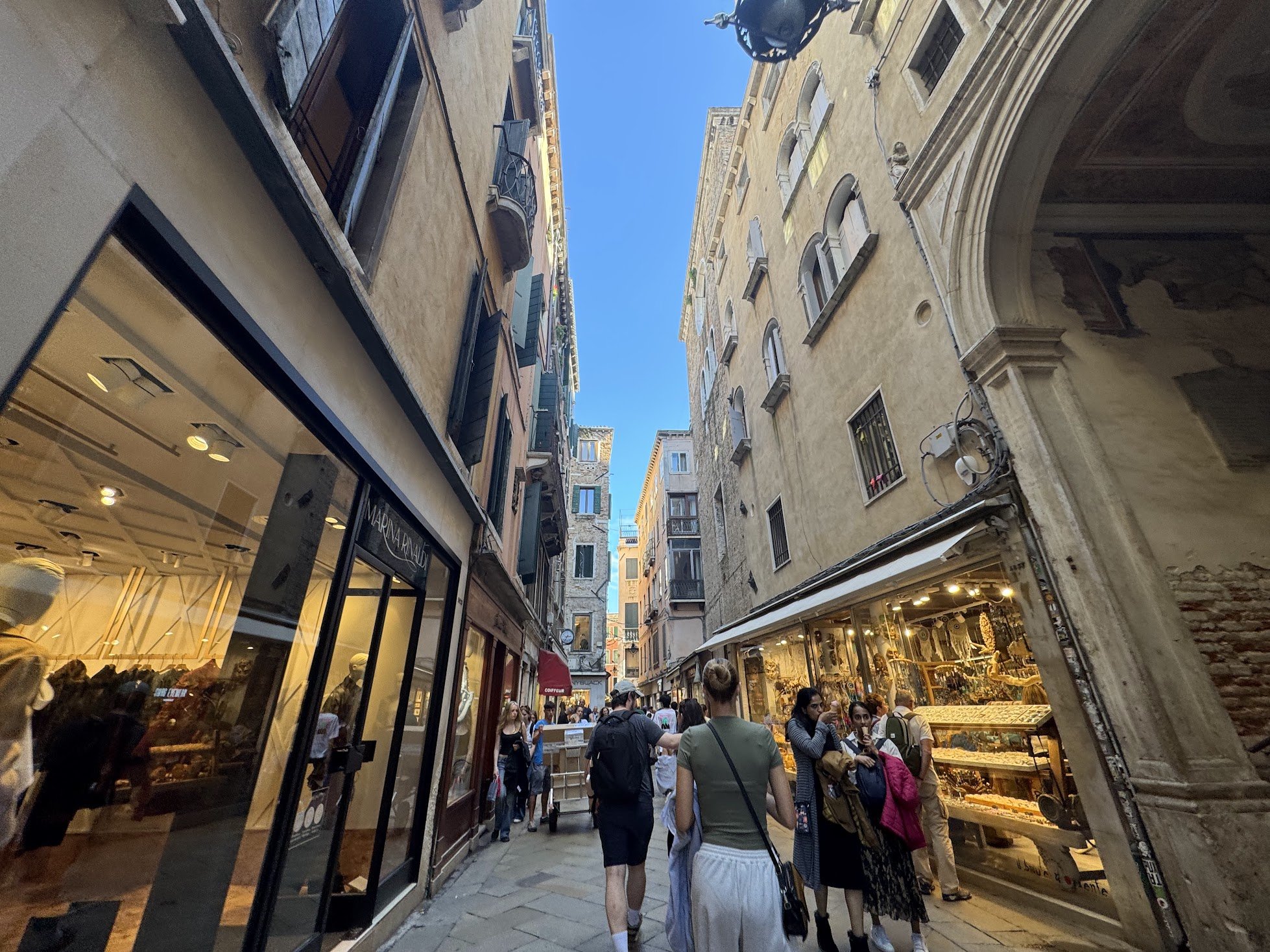A crowded pedestrian street lined with shops and cafes, with historic buildings and clear blue sky overhead. People are shopping and walking along the street.