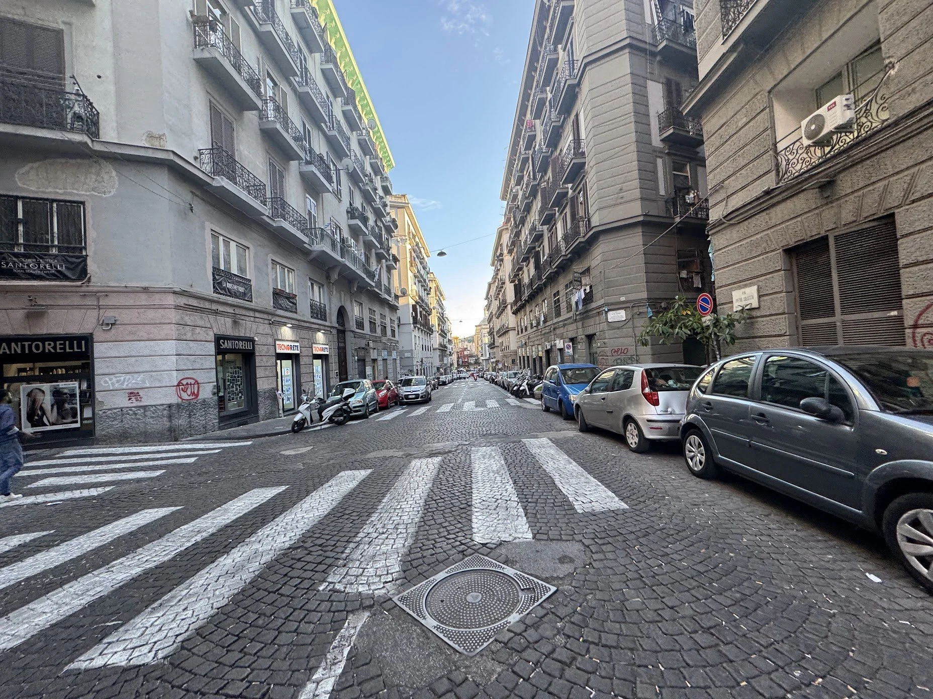 City street with cobblestone crosswalk, parked cars, tall buildings with balconies, and a clear blue sky.