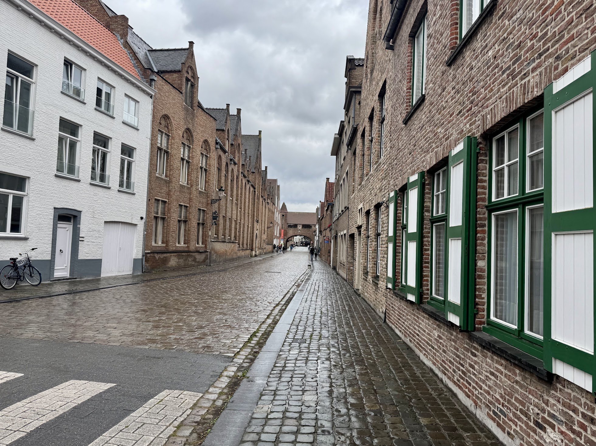 A narrow cobblestone street in a European city, lined with historic brick buildings. The buildings have windows with green shutters on one side and white and brick facades on the other. There are a few people walking in the distance, overcast sky, an
