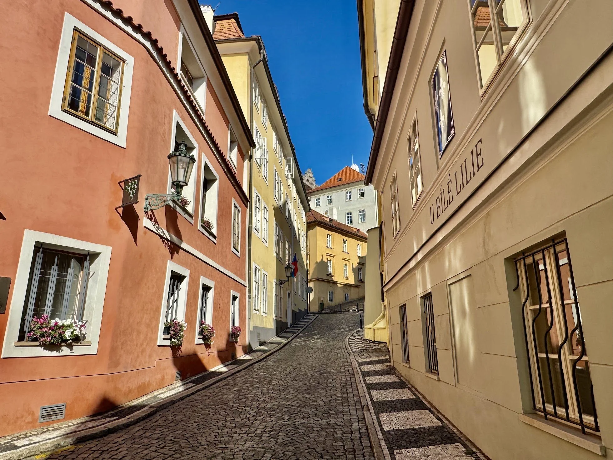 A narrow cobblestone street in a European city, lined with colorful buildings with flower boxes and street lamps, under a clear blue sky.